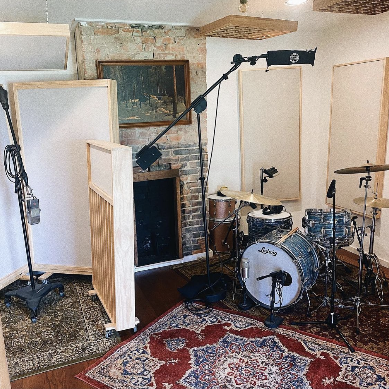 A music recording studio features a Ludwig drum set with microphones set up for recording, acoustic panels on the walls, a brick fireplace, and a painting of a forest scene hanging above the fireplace. The room has a patterned red and blue rug on the wooden floor.