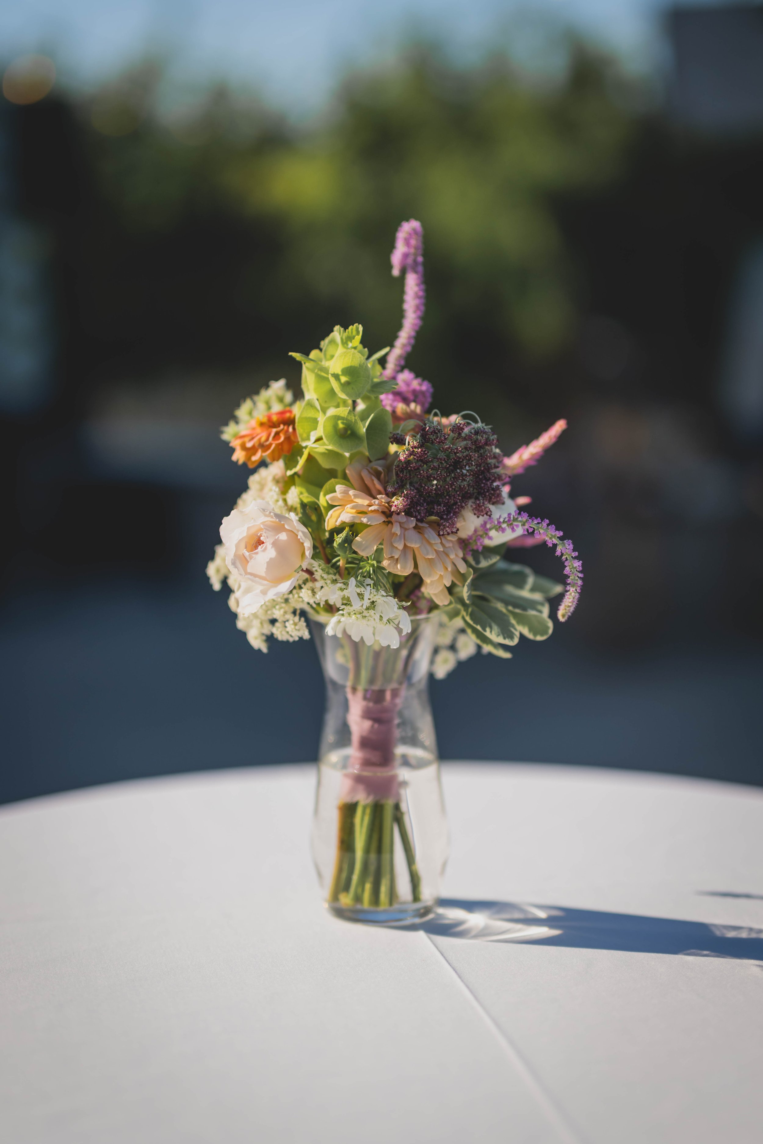 A bouquet of flowers, including pink, white, and purple blooms, in a clear glass vase on a white table, with a blurred green background.
