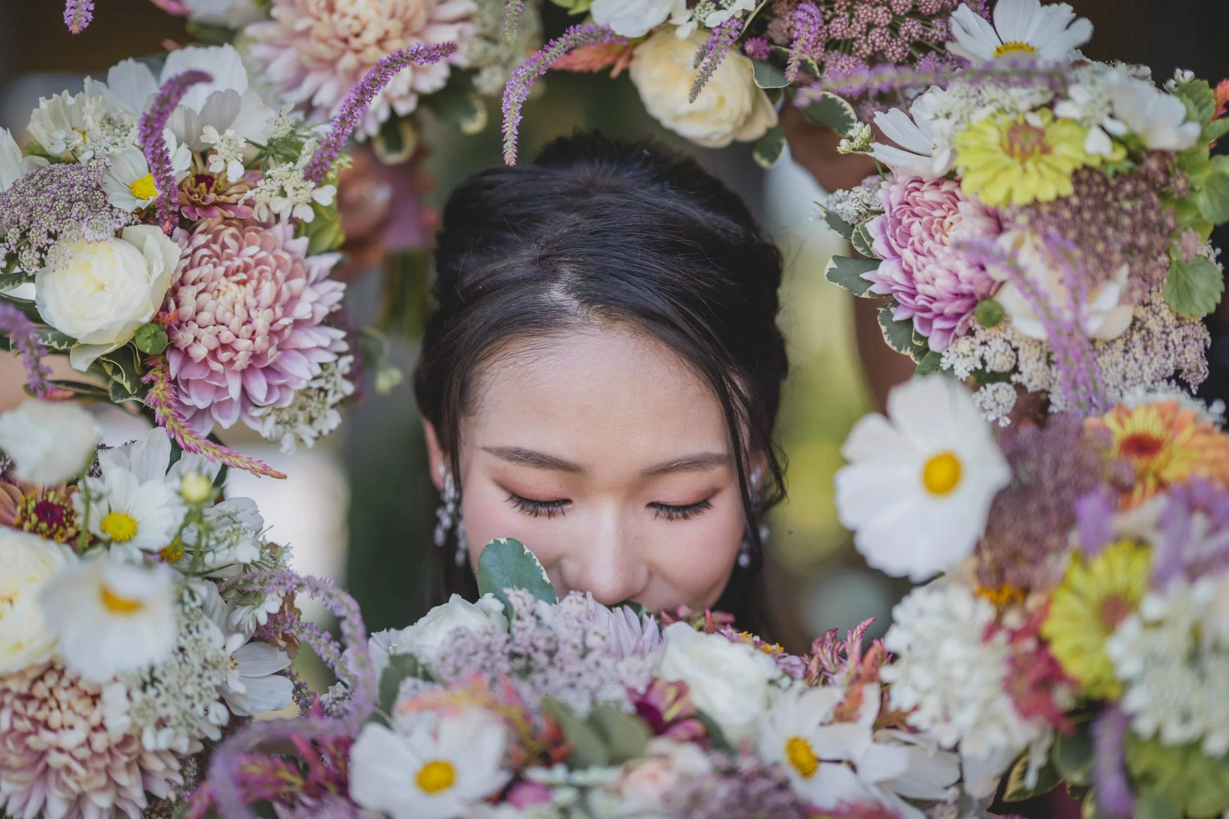 Woman surrounded by a wreath of colorful flowers, closing her eyes and smelling the bouquet in front of her.