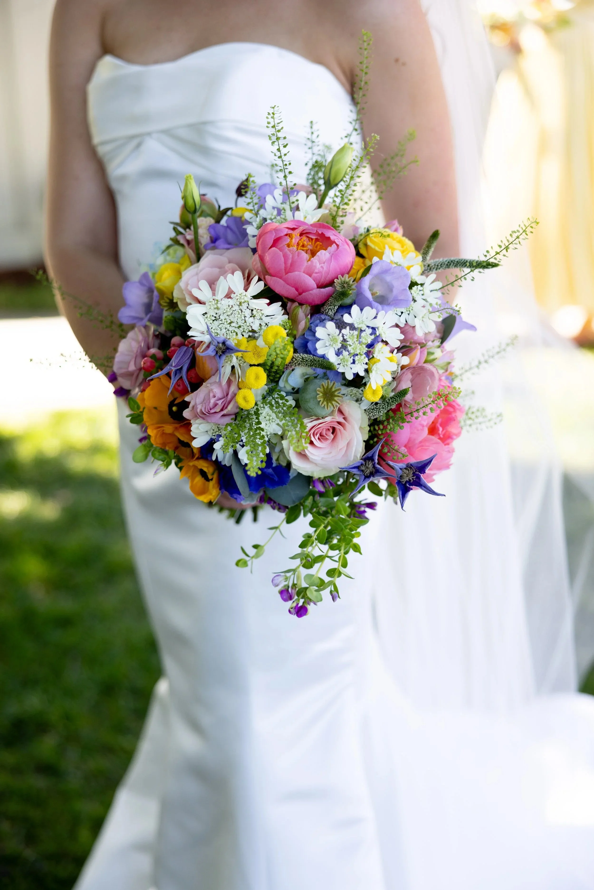 Bride holding a colorful bouquet of flowers against a white wedding dress background outdoors.