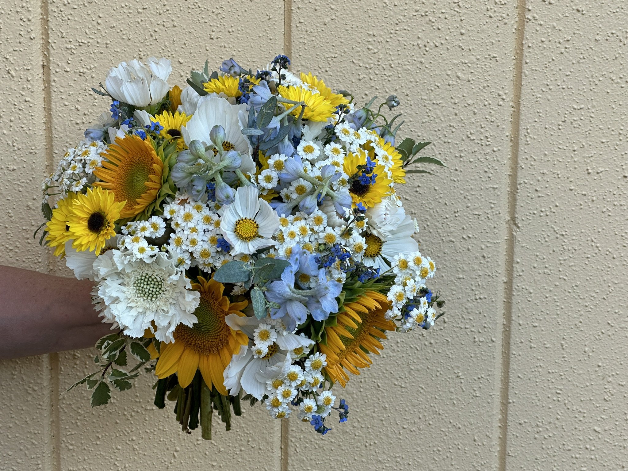 A bouquet of yellow and white flowers, including sunflowers and daisies, held against a textured beige wall.