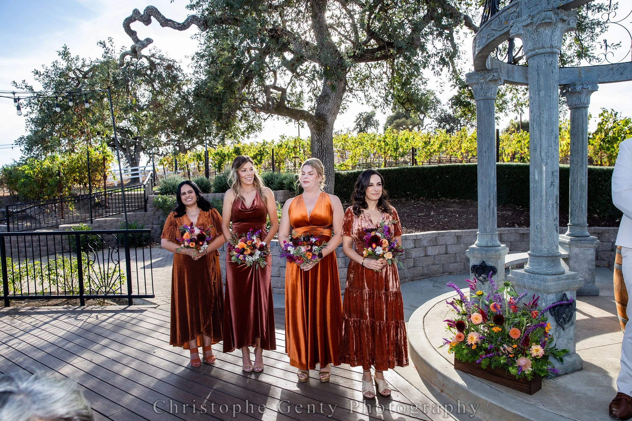 Four women in bridesmaid dresses holding bouquets, standing outdoors near a stone fountain and large tree, with vineyard in the background.