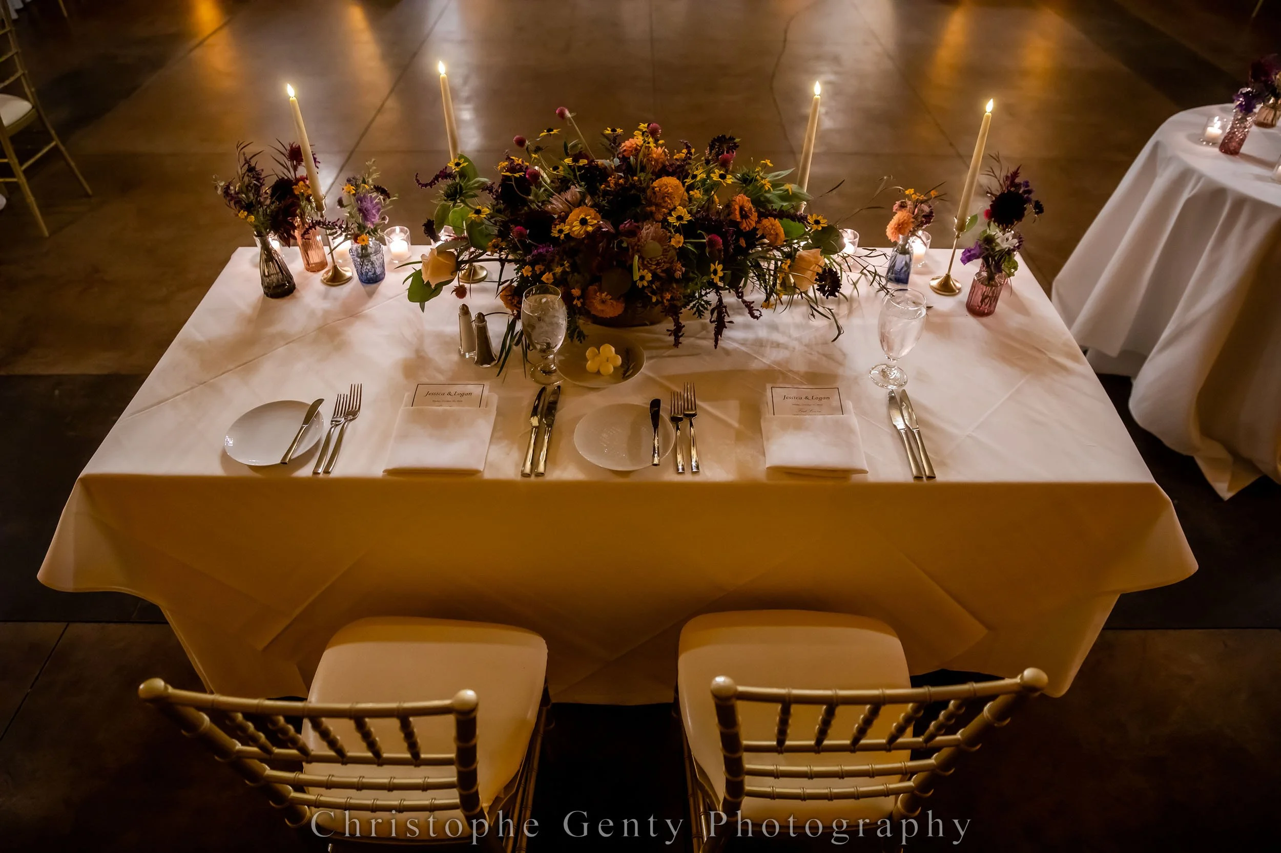 Elegant banquet table decorated with a large floral centerpiece, surrounded by candles, small flower arrangements in colorful vases, and set with white plates, silverware, and water glasses. The setting includes place cards and a candlelit ambiance.