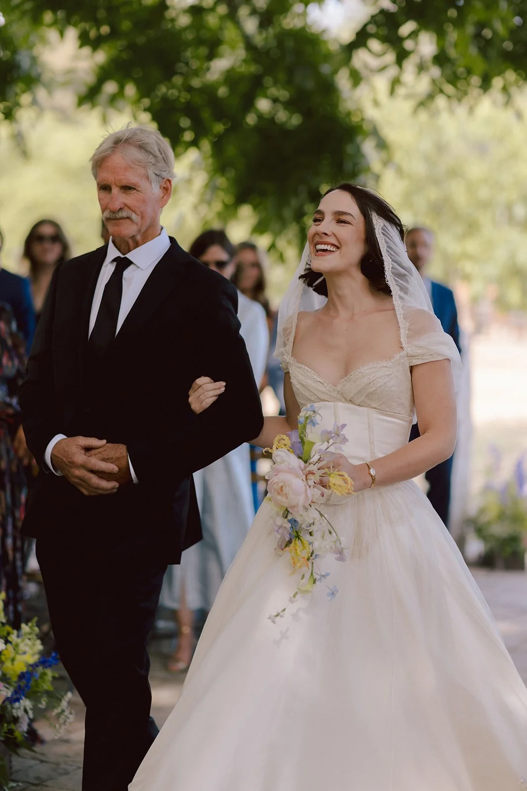 A bride in a white wedding dress and veil walking down the aisle with an older man in a black suit and tie, smiling and holding a bouquet of flowers, outdoors with trees and wedding guests in the background.