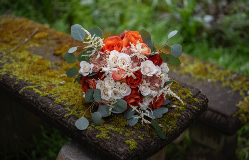 Bouquet with white, red, and orange flowers on mossy wooden bench.