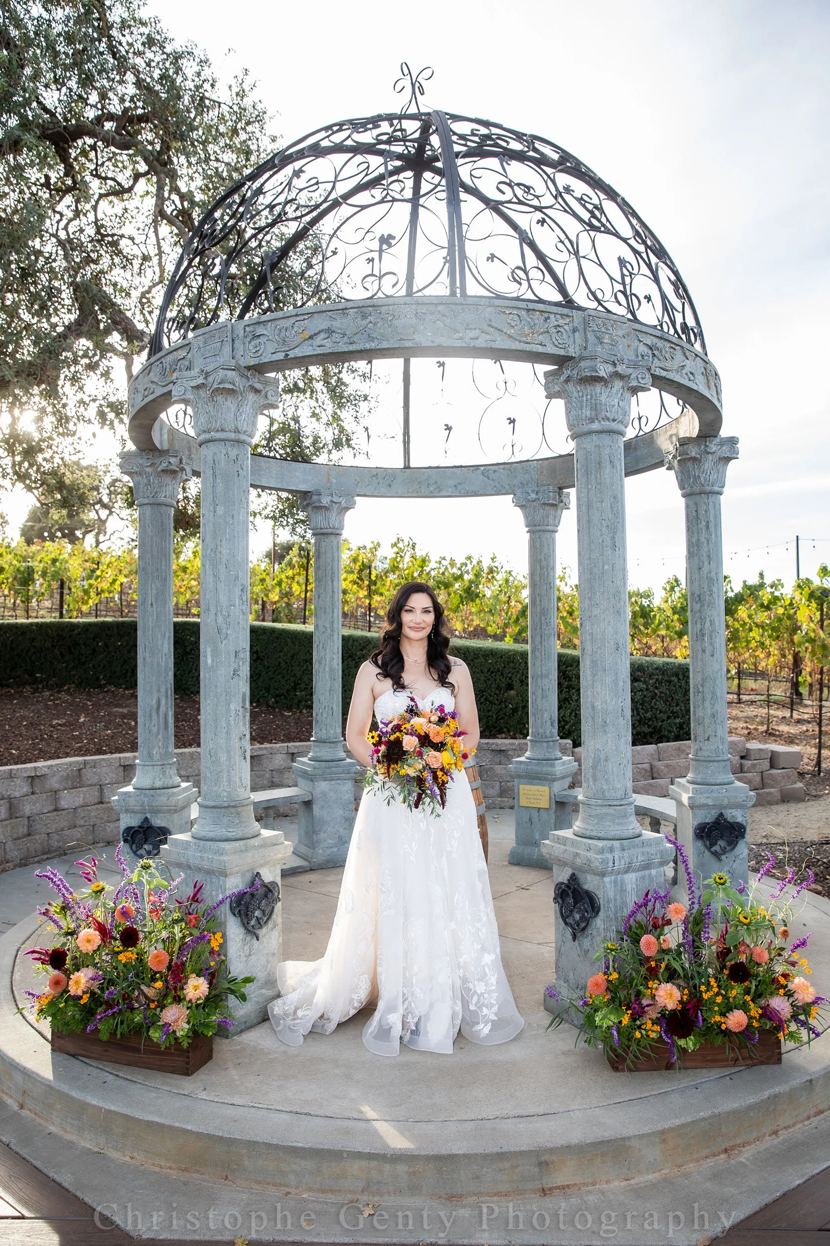 A woman in a white wedding dress holding a colorful bouquet standing under a decorative stone gazebo with pillars, surrounded by vibrant flower arrangements, outdoors during daytime.