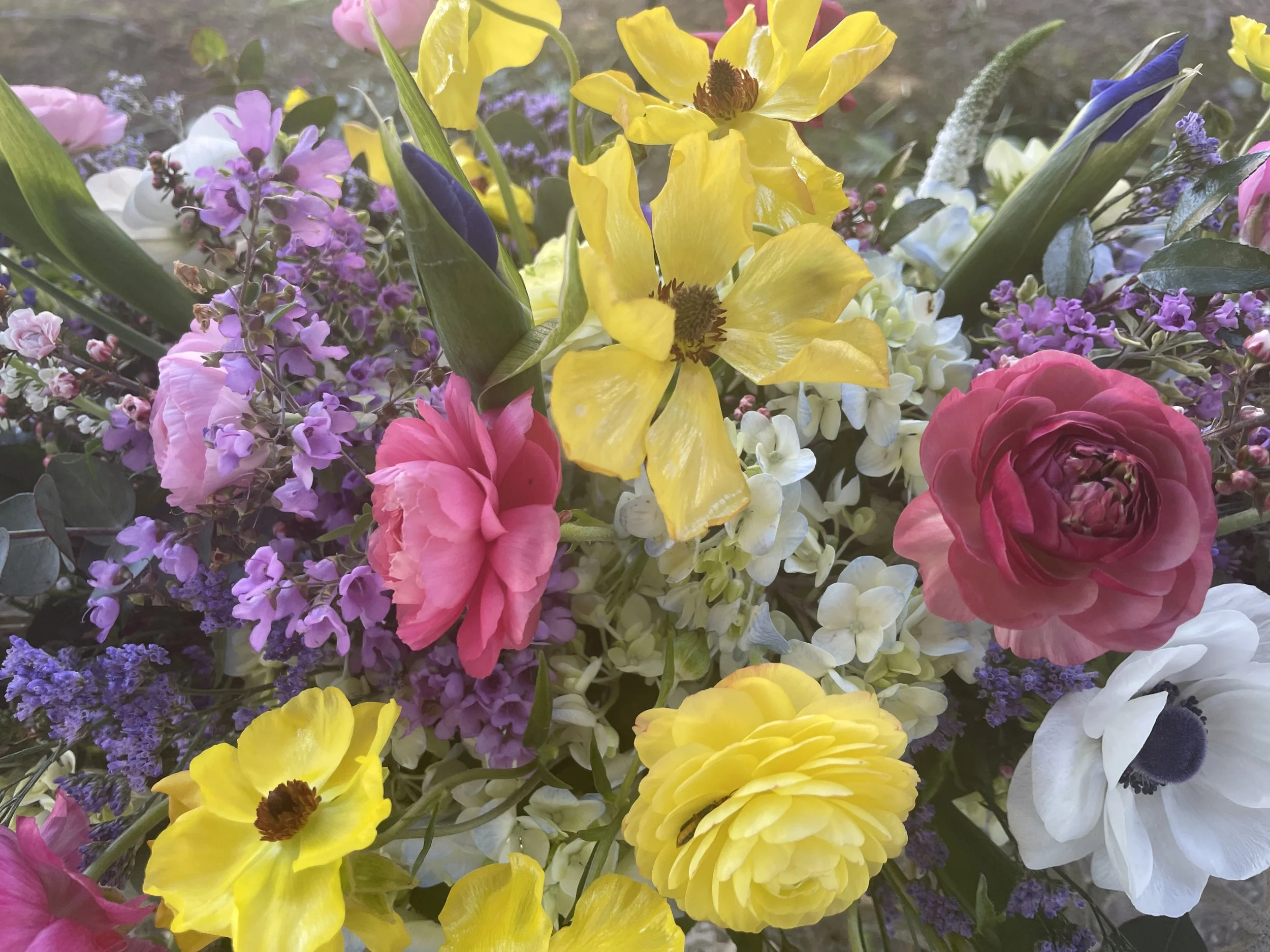 A colorful bouquet featuring a variety of flowers including yellow daffodils, pink and purple blooms, white and pink ranunculus, and other mixed blossoms.