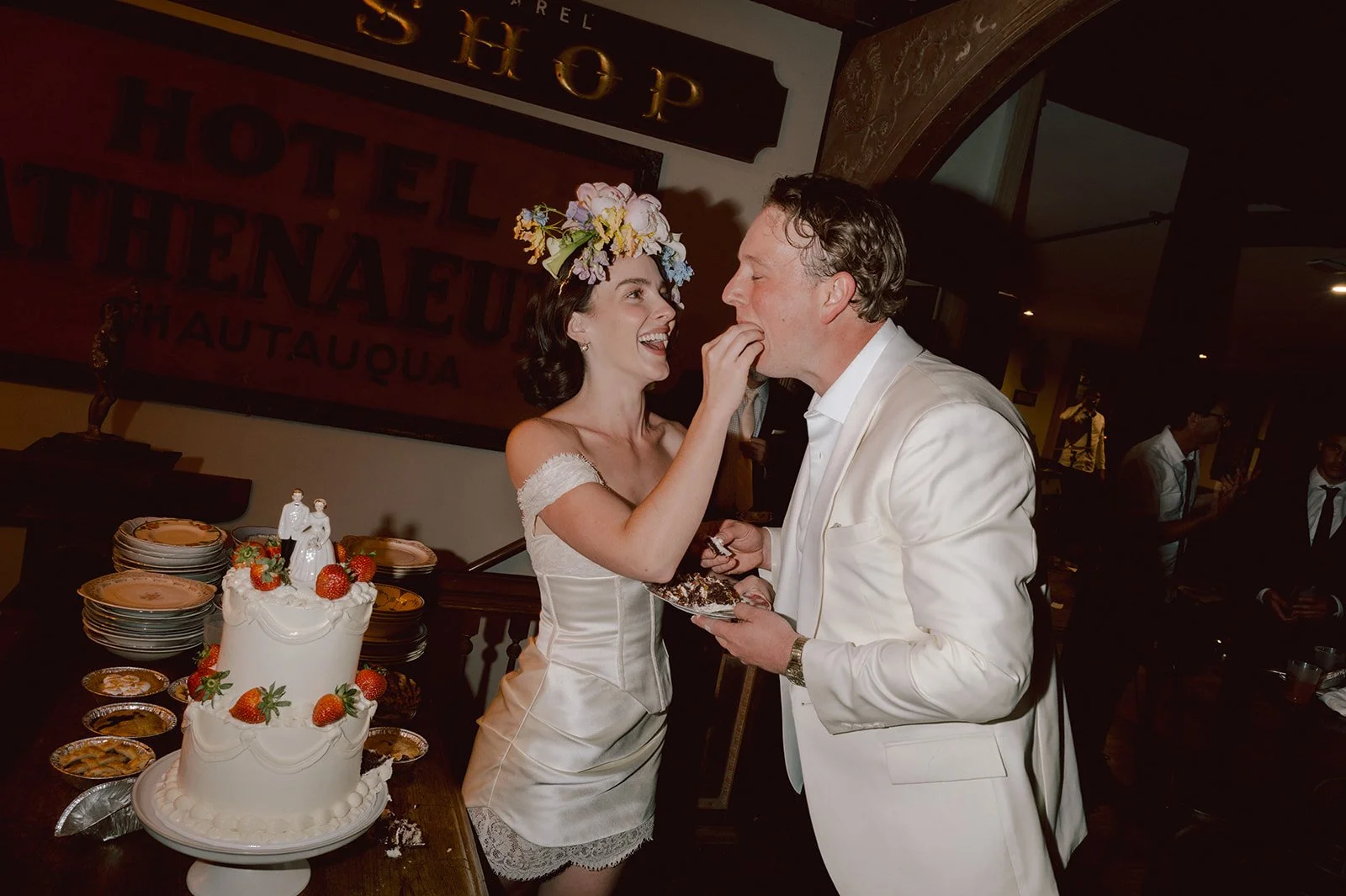 A bride with a floral headpiece touches a groom's nose as they smile at each other during their wedding reception. A wedding cake with strawberries is on the table in front of them, surrounded by plates and desserts. The background features a sign wi