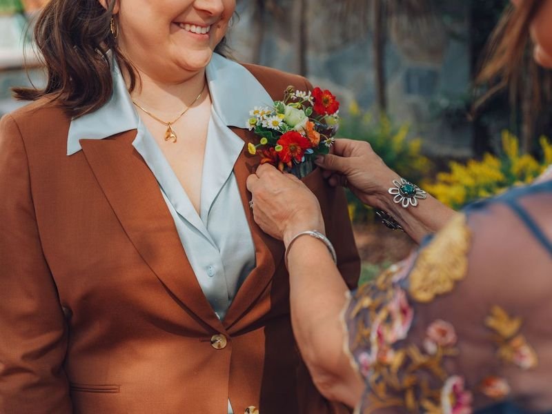 A woman in a brown blazer is receiving a boutonniere, a small flower arrangement, on her lapel from another woman during a celebration or ceremony.