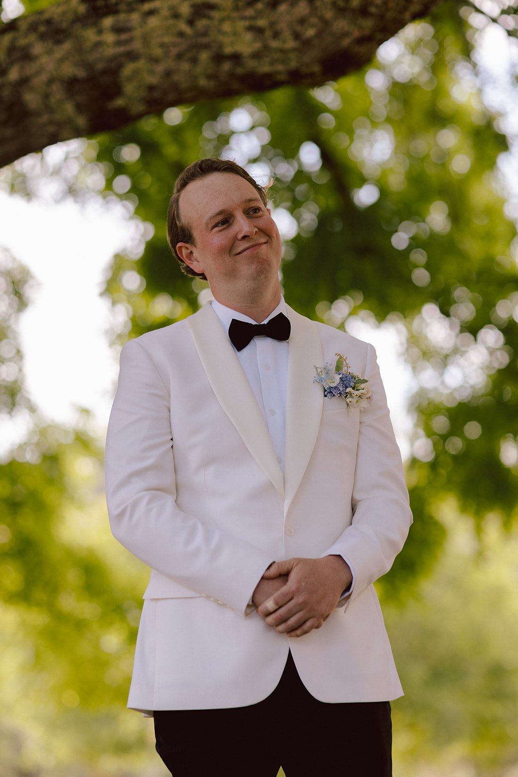 A man in a white tuxedo with a black bow tie, standing outdoors under a large tree with green leaves, smiling thoughtfully.