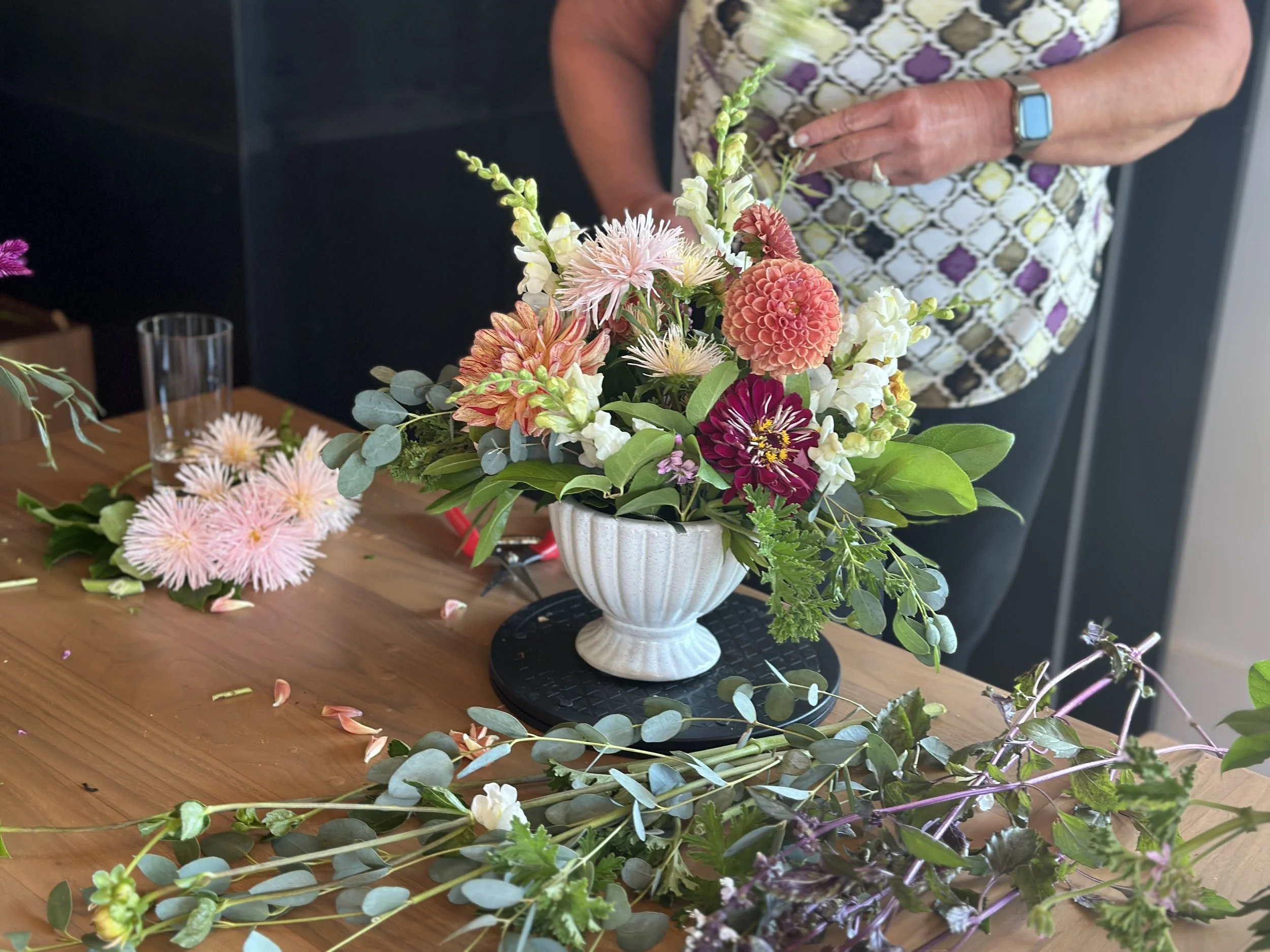 A person arranging a floral centerpiece on a wooden table. The arrangement includes a variety of flowers such as dahlias and chrysanthemums, with greenery and leaves, in a white vase. Additional flower stems and leaves are scattered on the table.