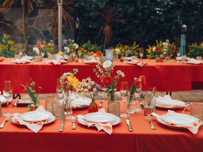 A decorated outdoor banquet table with floral centerpieces, white plates with peach napkins, and glassware, set against a garden backdrop.