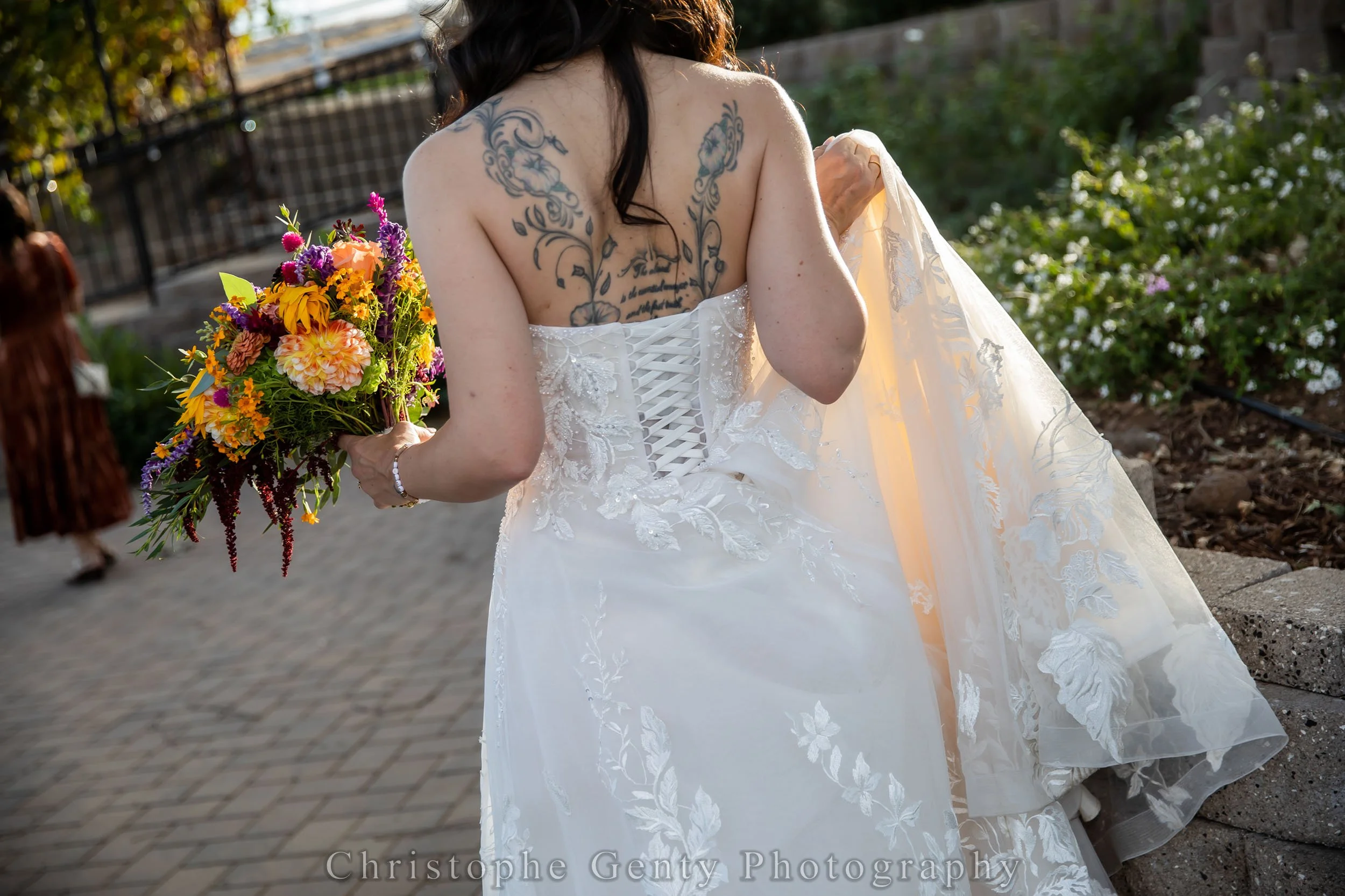 A bride dressed in a white wedding gown holding a colorful bouquet of flowers, standing outdoors with greenery and a paved path surrounding her.