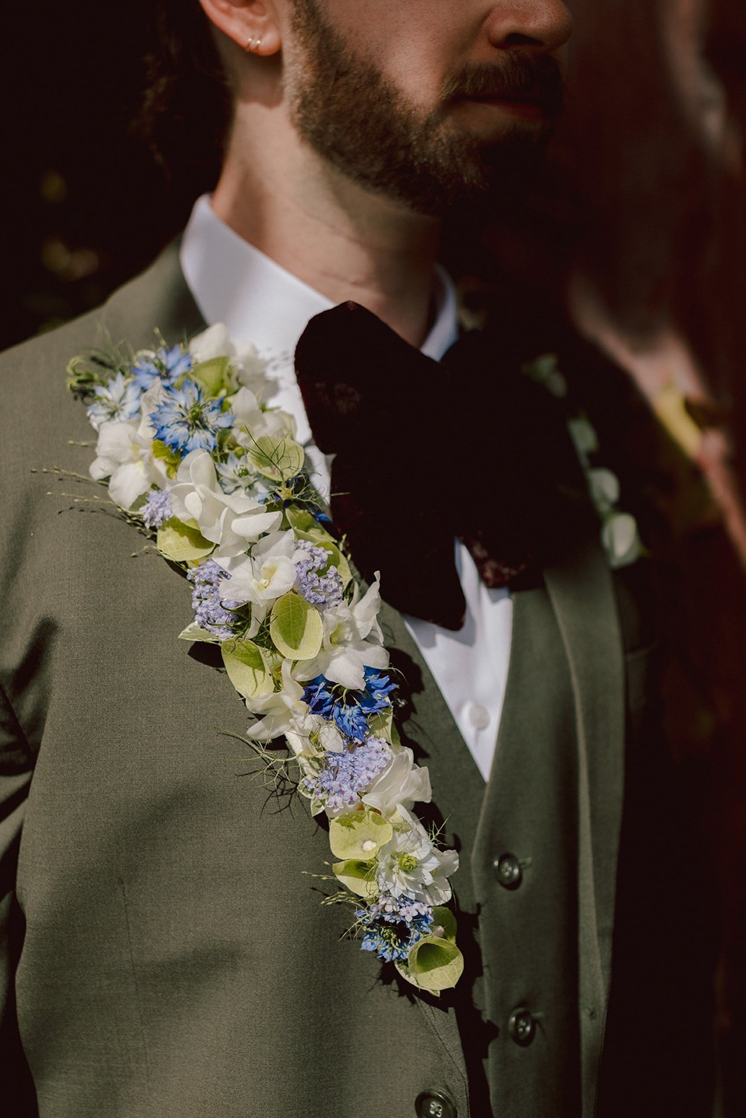 Close-up of a man wearing a green suit and black bow tie, with a floral arrangement on his lapel, including white, blue, and purple flowers and green leaves.