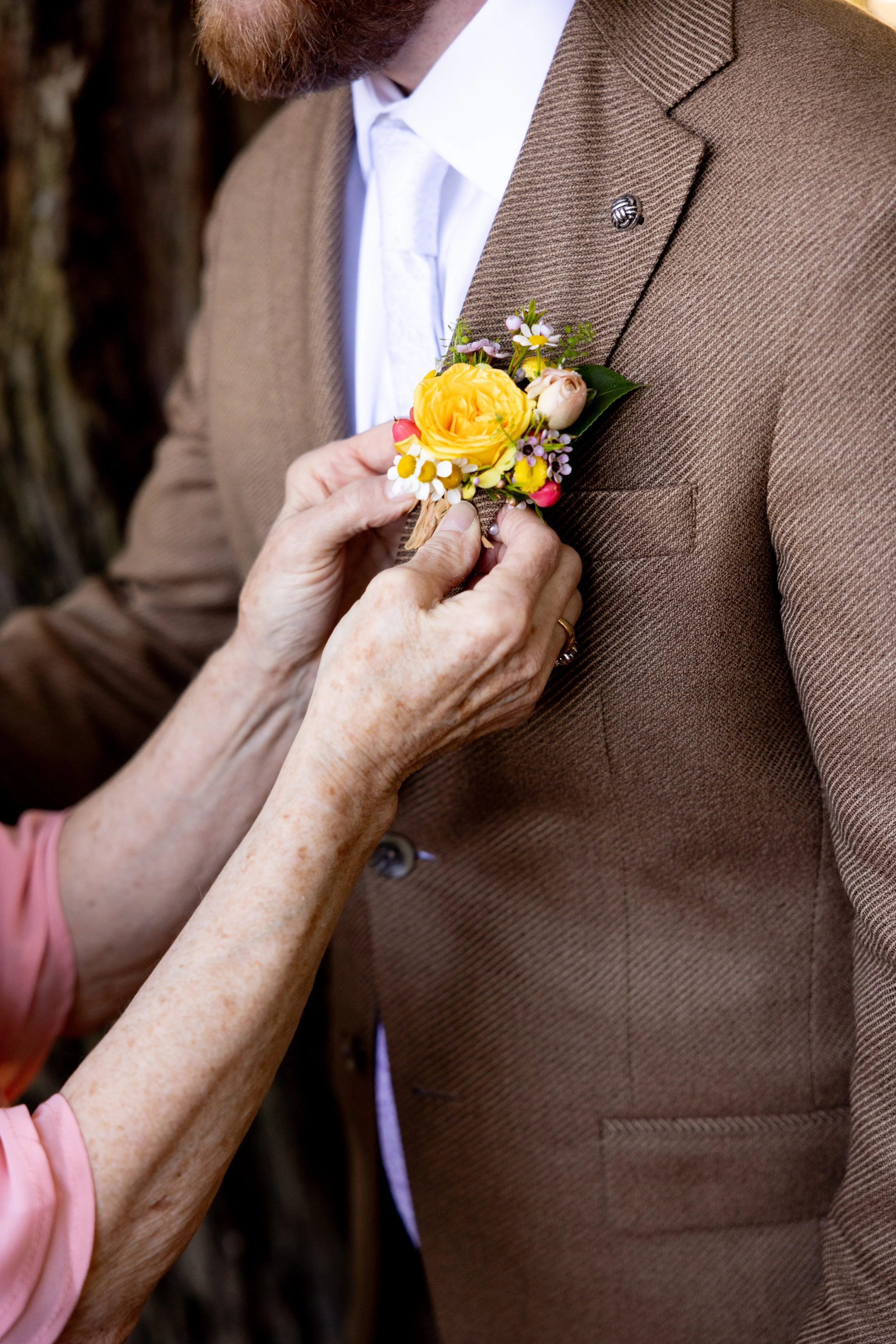 Someone is pinning a colorful boutonniere on a man's brown blazer jacket.