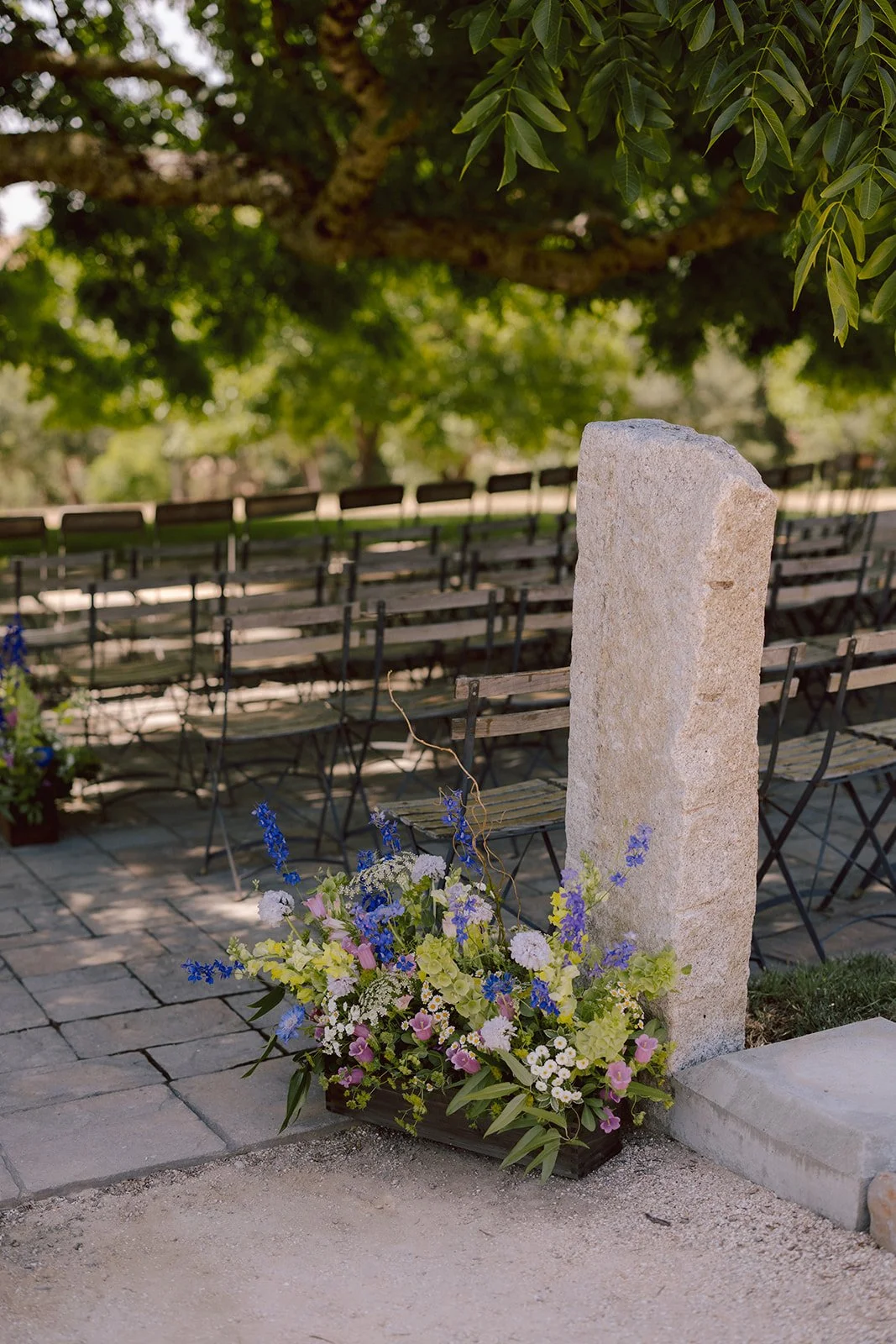 A memorial site with a stone marker and a flower arrangement in a black container at the base, surrounded by trees and benches in the background.