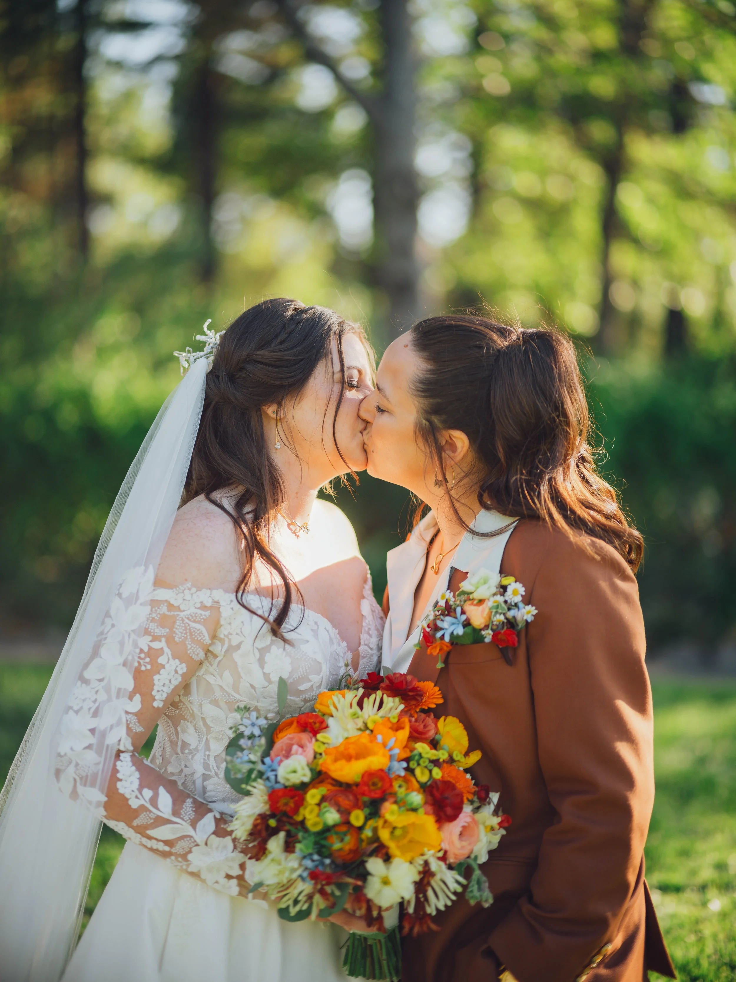 Two women, one in a wedding dress with lace details and a veil, and the other in a brown blazer with a flower pin, sharing a kiss with a bouquet of colorful flowers, outdoors in a green, wooded area.