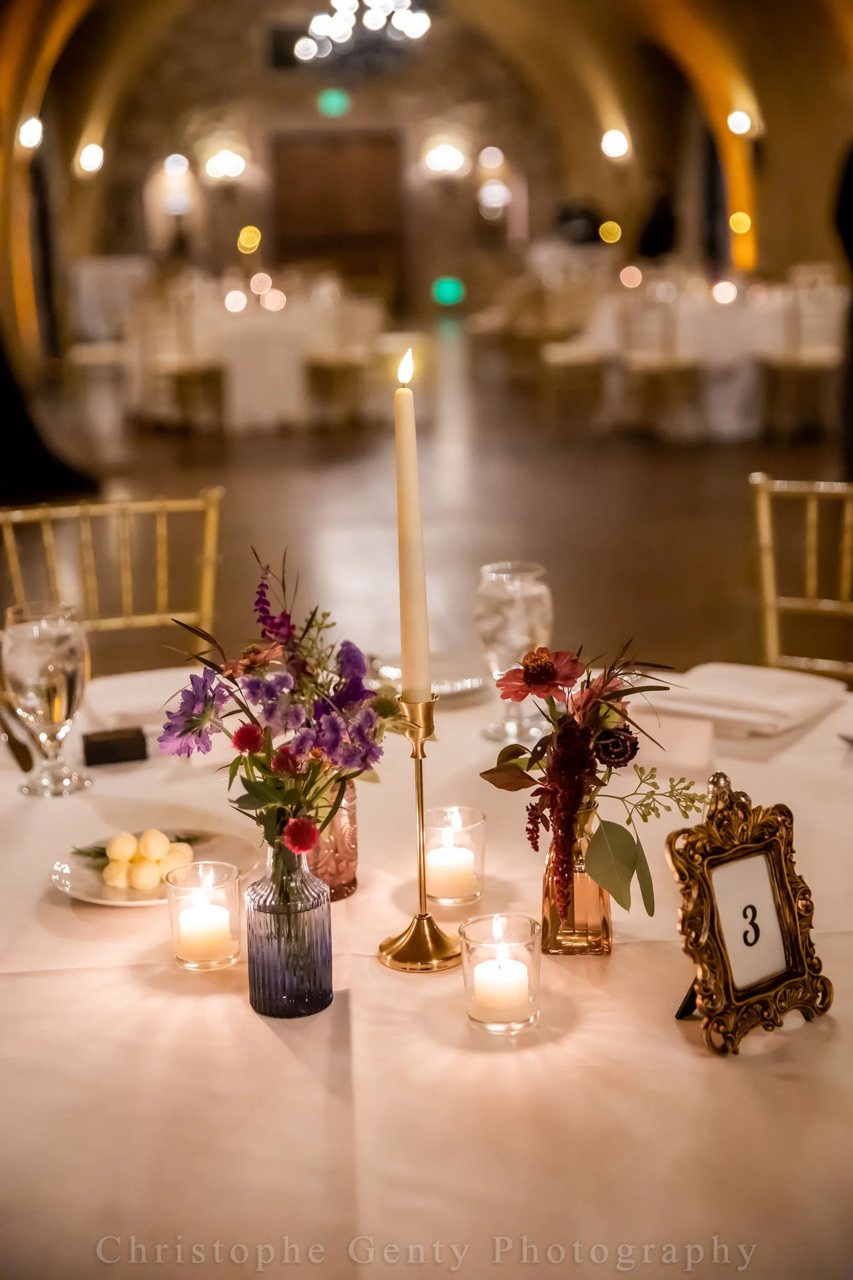 Elegant wedding reception table with lit candles, floral centerpieces, and a gold table number frame in a dimly lit venue with arched ceiling.
