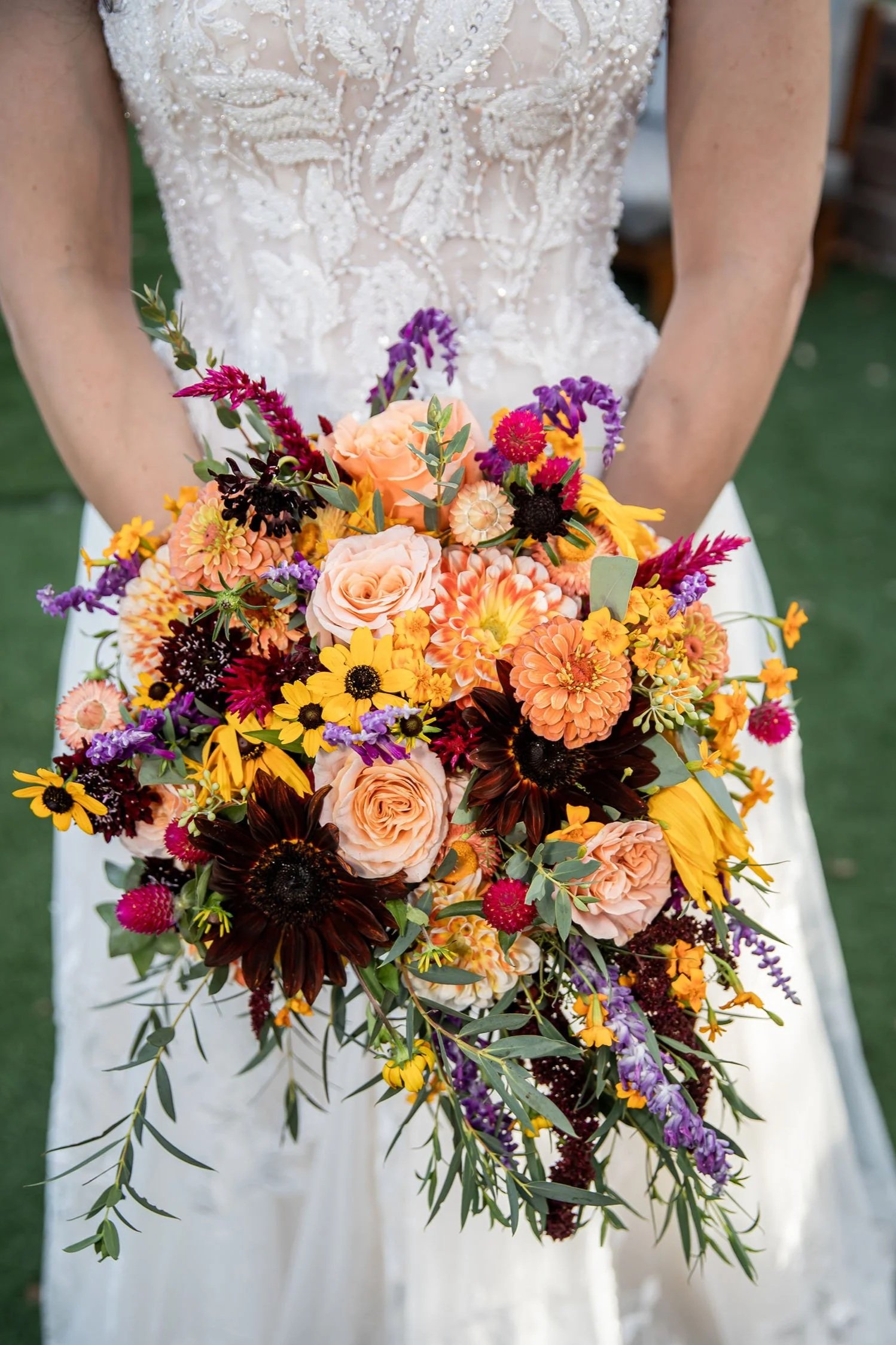 Bride holding a colorful seasonal wedding bouquet featuring roses, zinnias, sunflowers, celosia, and locally grown flowers.