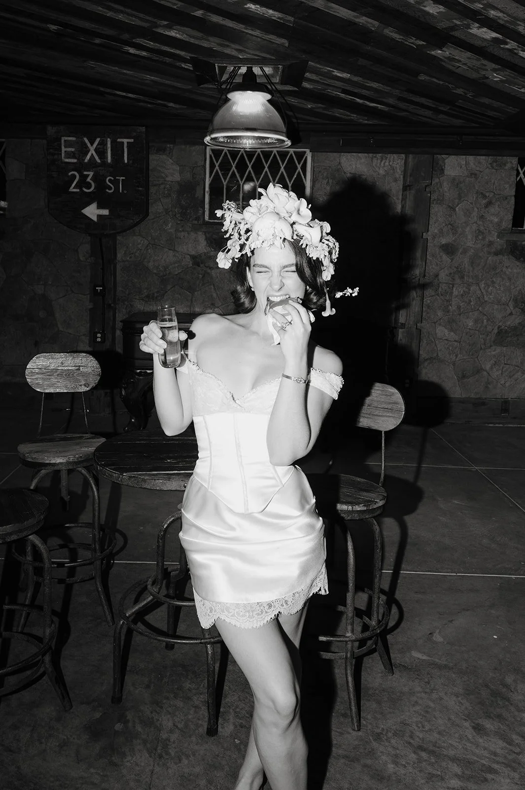A woman in a white dress with lace details, wearing a floral headpiece, is laughing and eating while holding a glass, in a rustic indoor setting with wooden chairs and a stone wall.