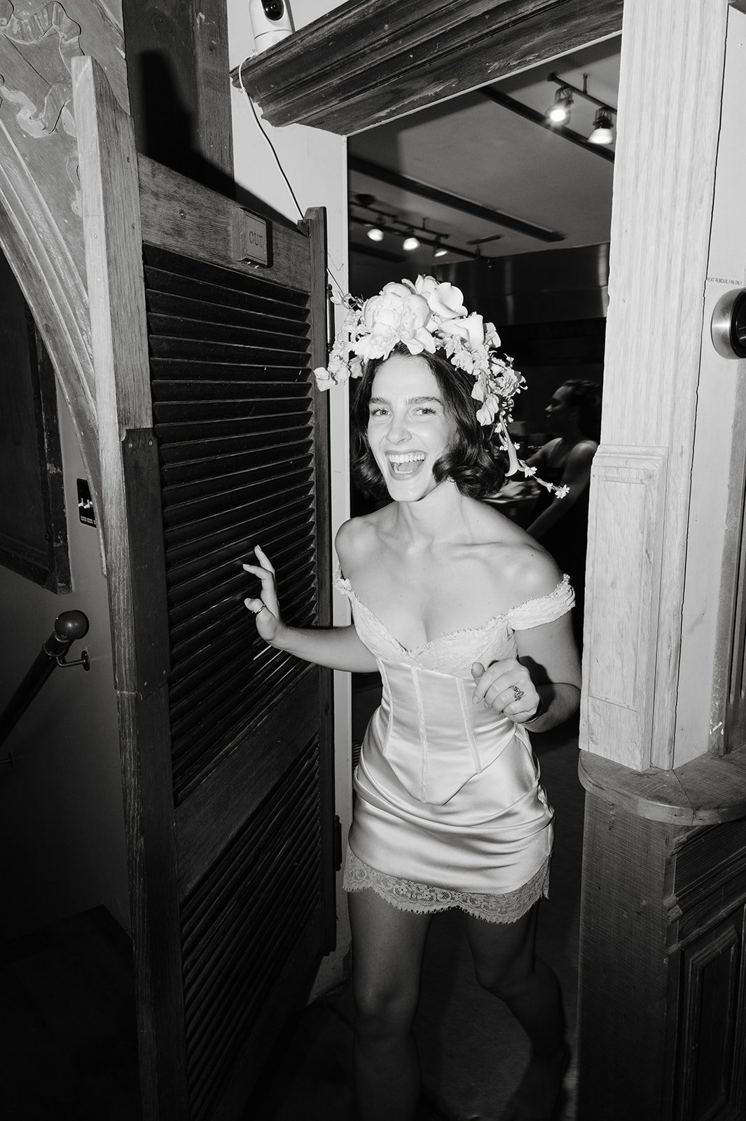 A woman in a satin off-the-shoulder dress with lace trim, wearing a large floral crown, smiling and playfully peeking out from behind a wooden shutter door in a rustic setting.