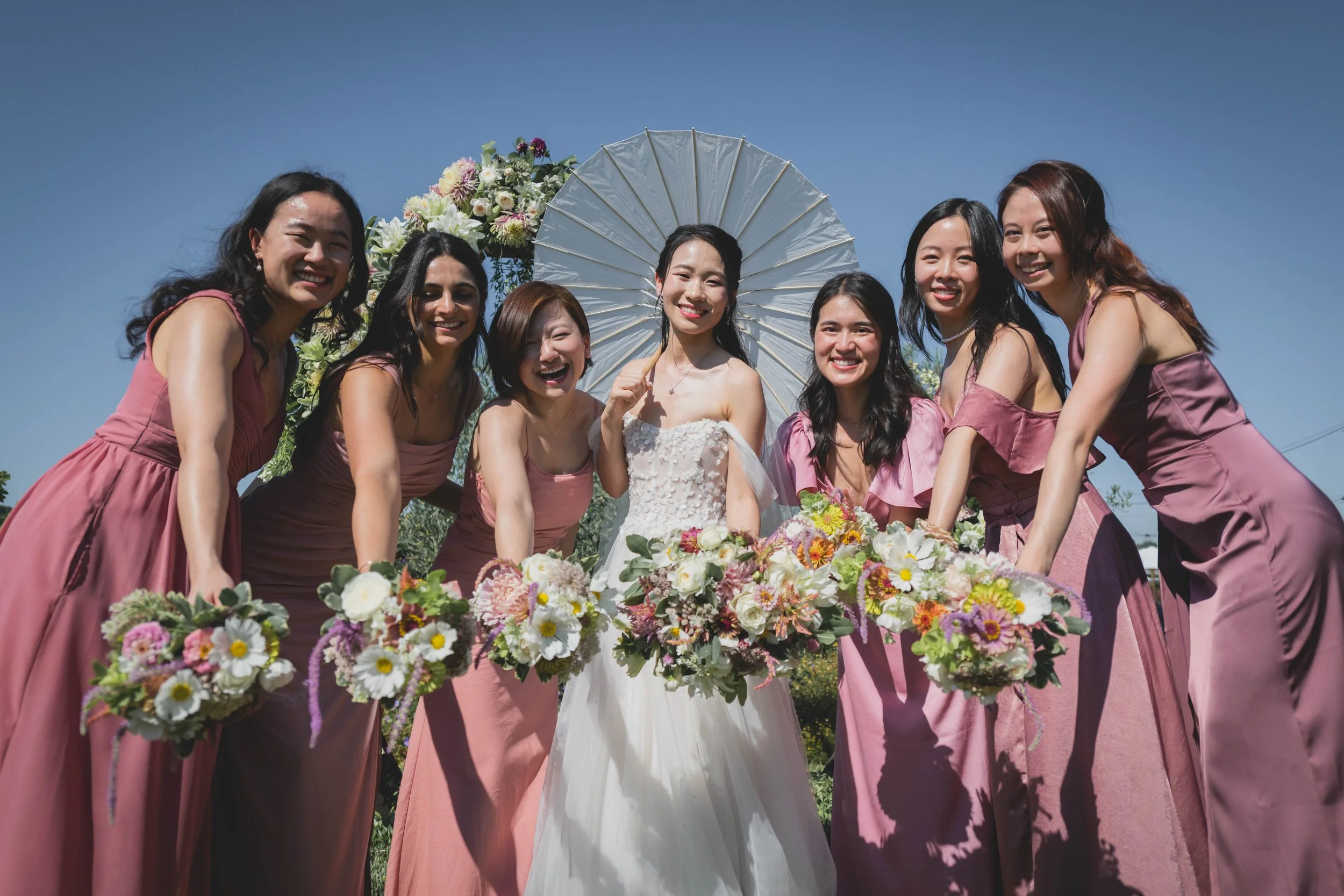 bride holding a white parasol and six bridesmaids wearing dusty rose gowns