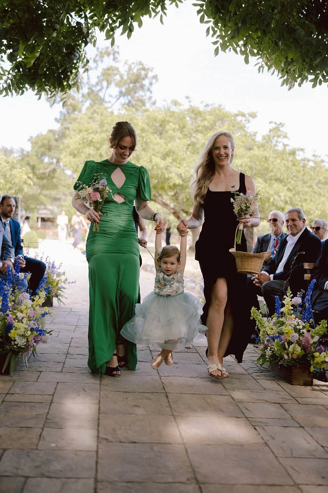 Two women, one in a green dress and one in a black dress, are walking down the aisle holding a young girl's hands. The girl, in a floral top and white tulle skirt, is being lifted slightly off the ground. They are outdoors with trees and seated guest