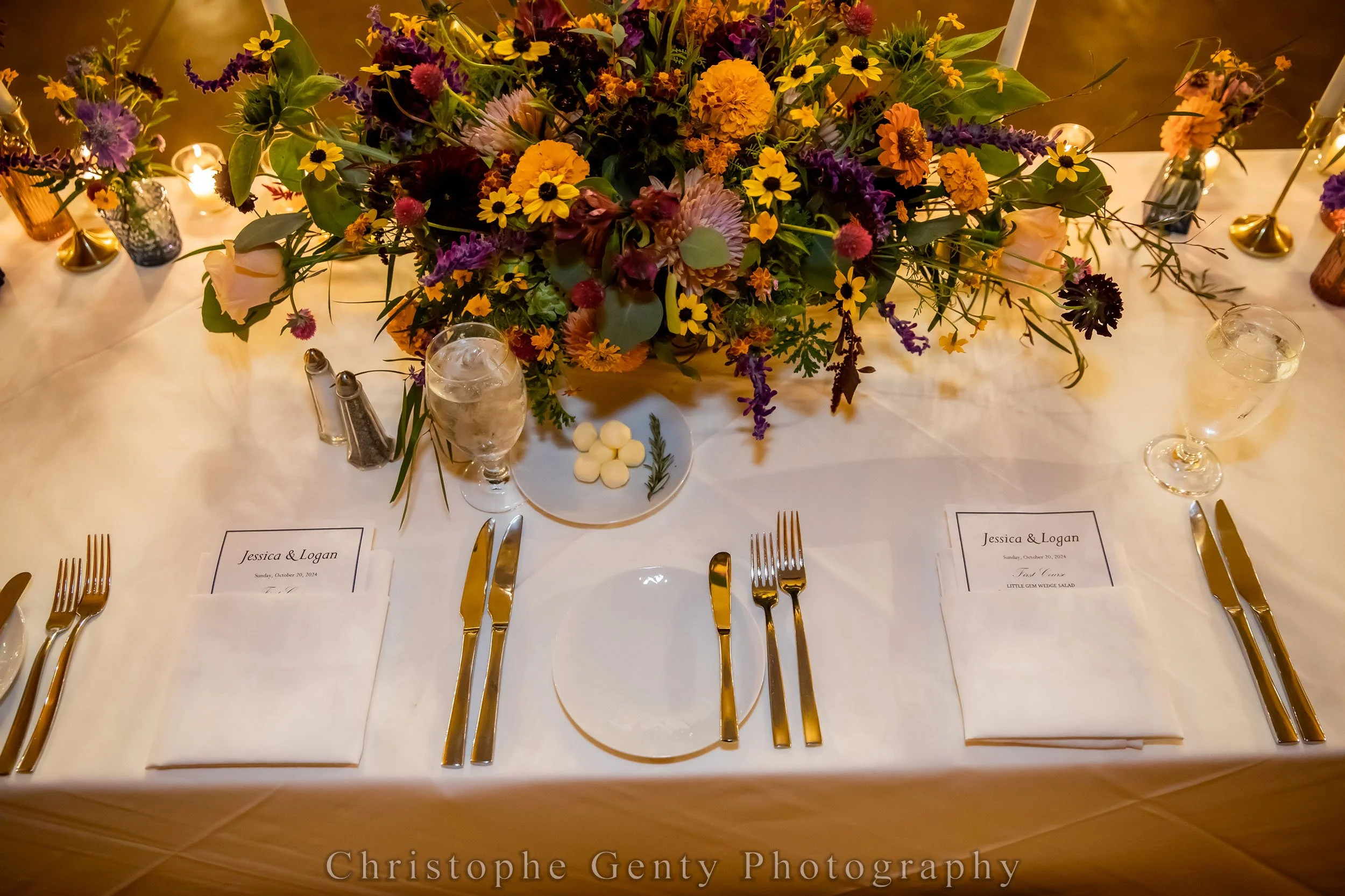 A decorated table setting with a large floral arrangement of purple, yellow, orange, and pink flowers taking center stage. The table includes white tablecloths, neatly folded napkins with printed menus, and gold-colored cutlery. There are water glass