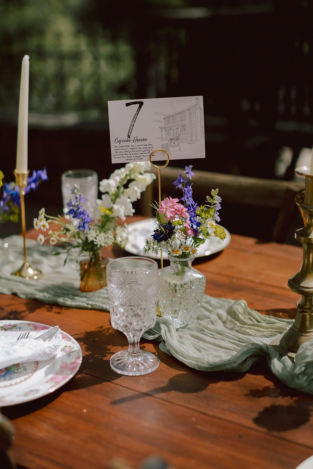 A rustic dining table setup with floral centerpieces in glass jars, a table number card, crystal glasses, floral patterned plates, and brass candlesticks with candles.