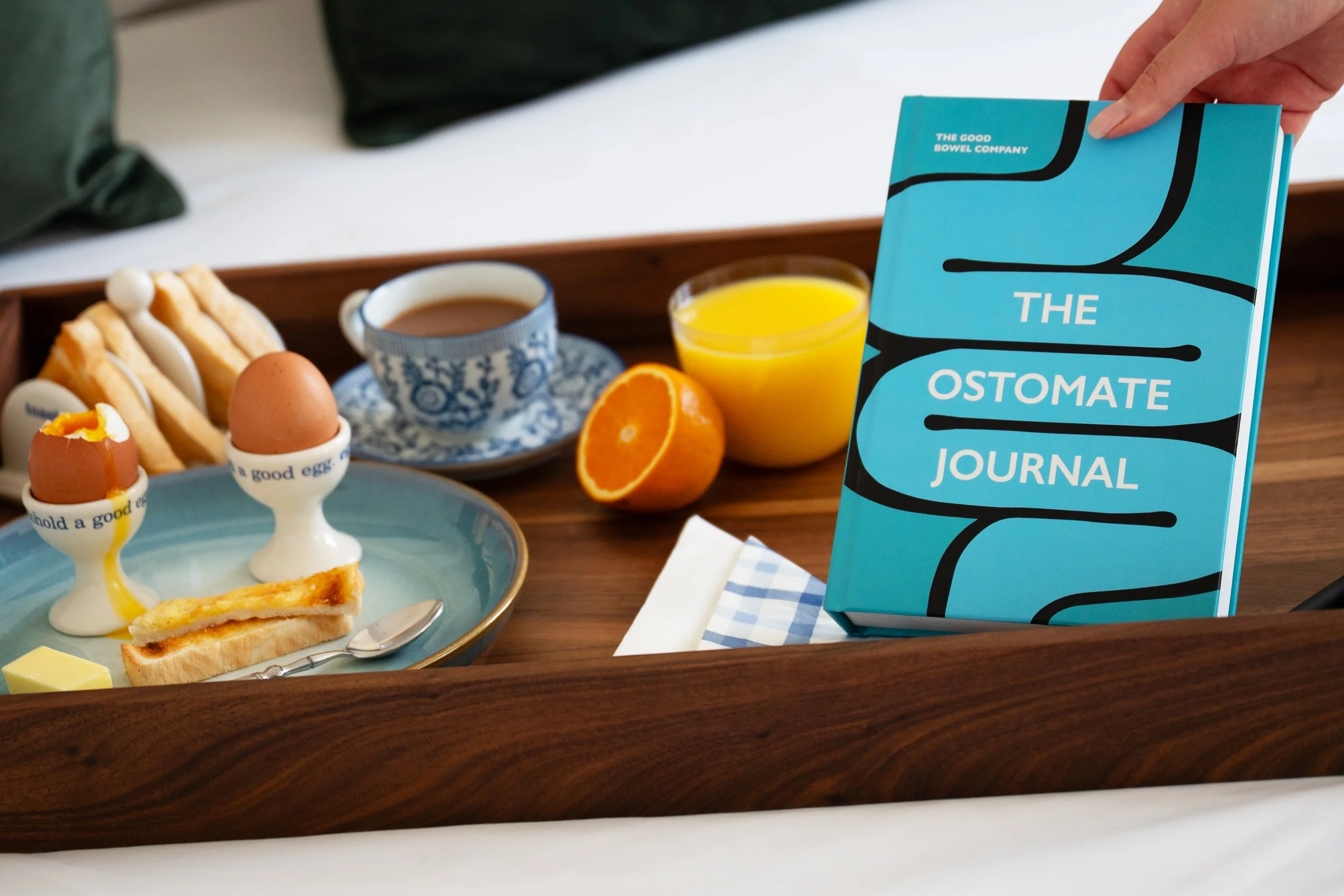 Breakfast on a wooden tray including toast with eggs, a cup of tea, a glass of orange juice, half an orange, and a blue journal titled 'The Ostomate Journal' being held up.