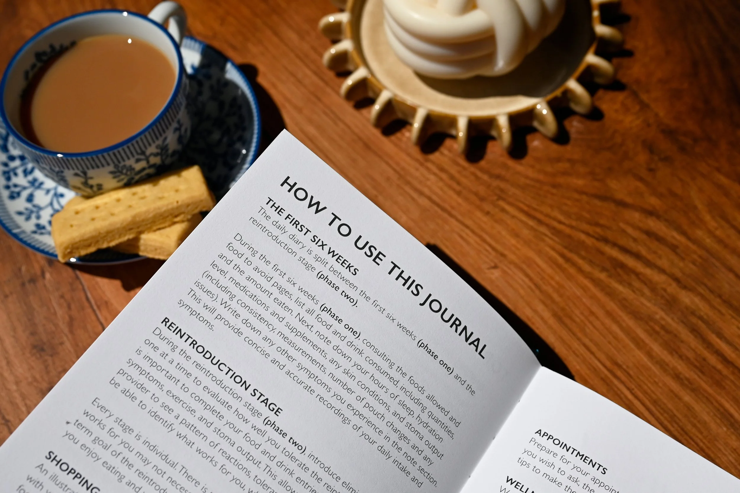 A journal with instructions for how to use The Ostomate Journal placed on a wooden table. To the left, there is a cup of tea with a biscuit beside it