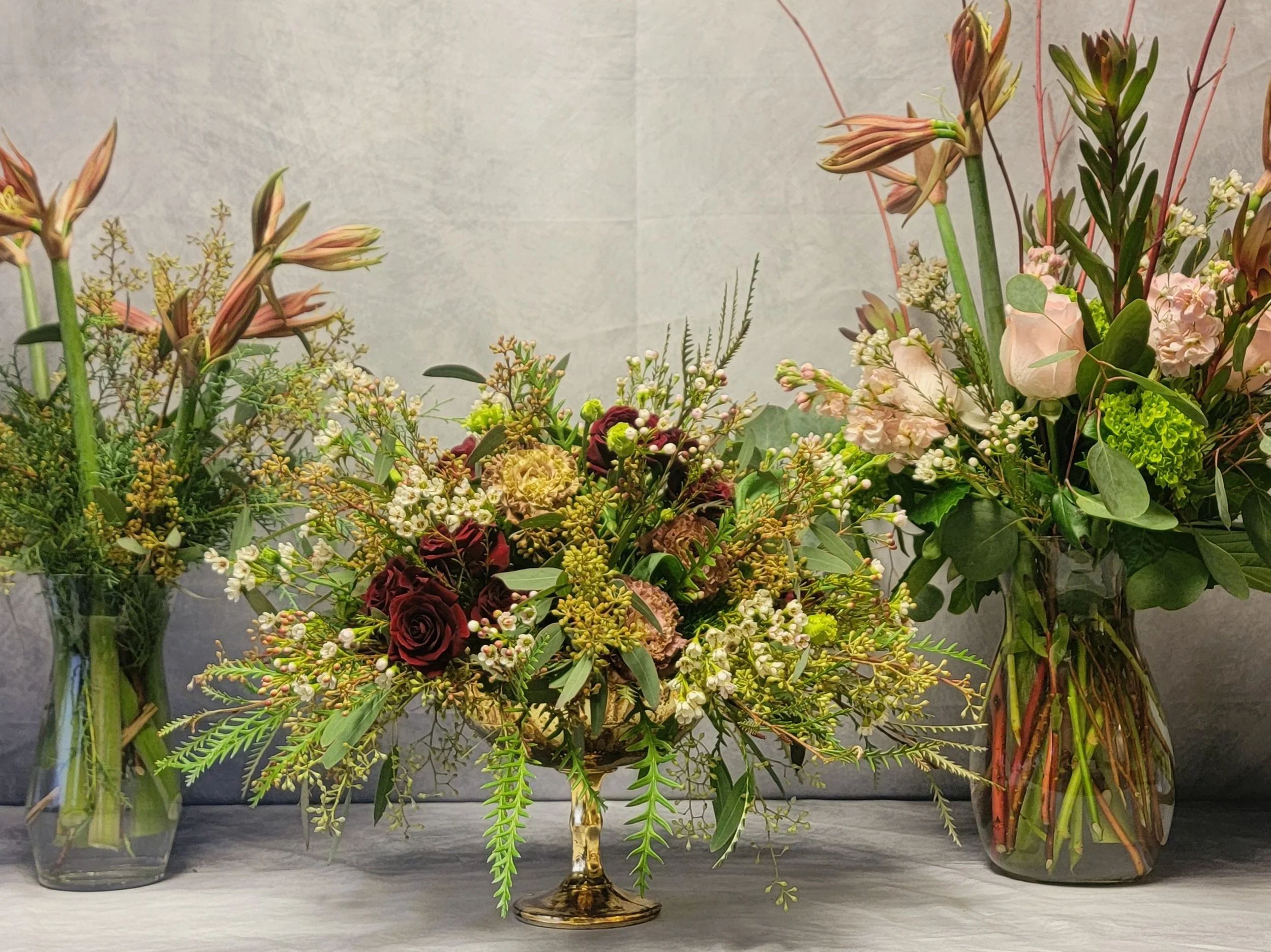 Arrangement of three floral vases with various flowers and greenery, including roses and lilies, placed on a flat surface against a plain background.