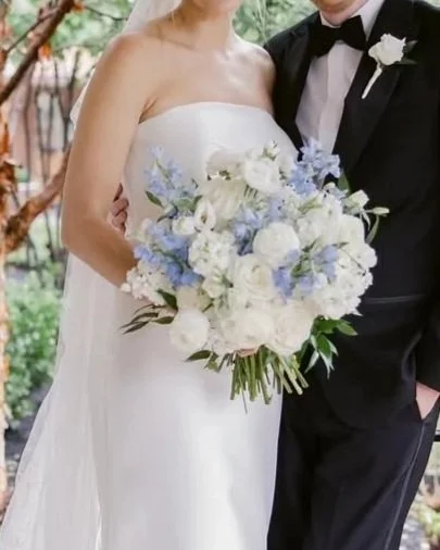 Bride in a white strapless wedding dress holding a large bouquet of white and light blue flowers, standing next to groom in a black tuxedo with a white shirt and black bow tie, outdoors with trees in the background.