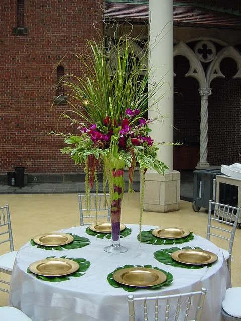 A round banquet table with a white tablecloth, six gold dinner plates, and green leaves under each plate. A tall floral centerpiece with green grasses and purple and pink flowers.