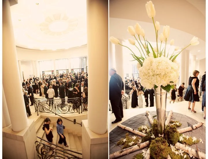 A large indoor gathering or event with many people dressed in formal attire, featuring elegant decor, including a prominent floral arrangement in a tall vase with white flowers and greenery, and a spacious, well-lit hall with high ceilings and curved