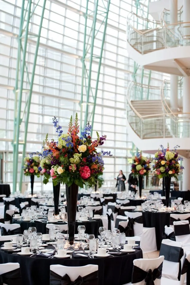 Elegant banquet hall with round tables covered in black tablecloths, each with tall floral centerpieces, glassware, and place settings, inside a modern glass atrium with curved staircases.