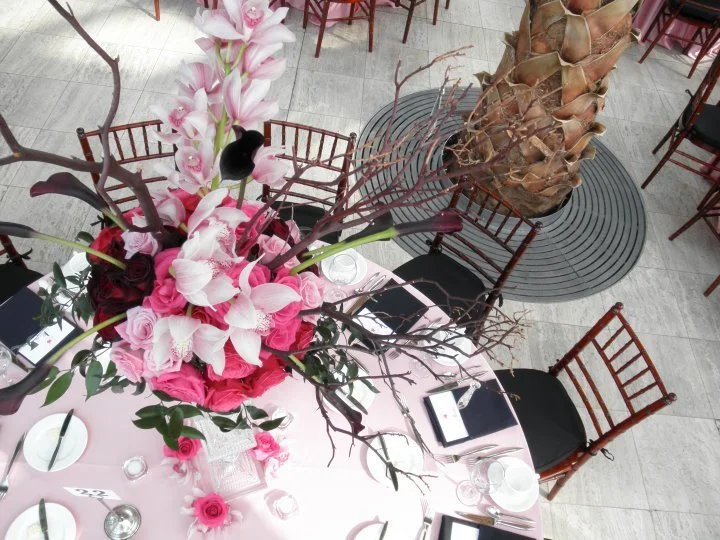 Overhead view of a table with a large pink floral centerpiece, place settings with glasses and black menus, surrounded by wooden chairs, with potted plants in the background.