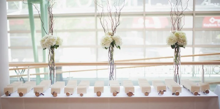 Table with rolled-up certificates or scrolls and floral arrangements in vases, set up indoors near large windows.
