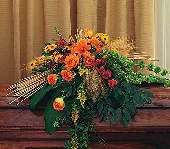 A large floral arrangement with orange, yellow, pink, and red flowers, green leaves, and wheat stalks on a wooden surface in front of beige curtains.