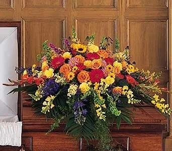 Large floral arrangement of yellow, pink, purple, and orange flowers on a wooden surface against wood paneling.
