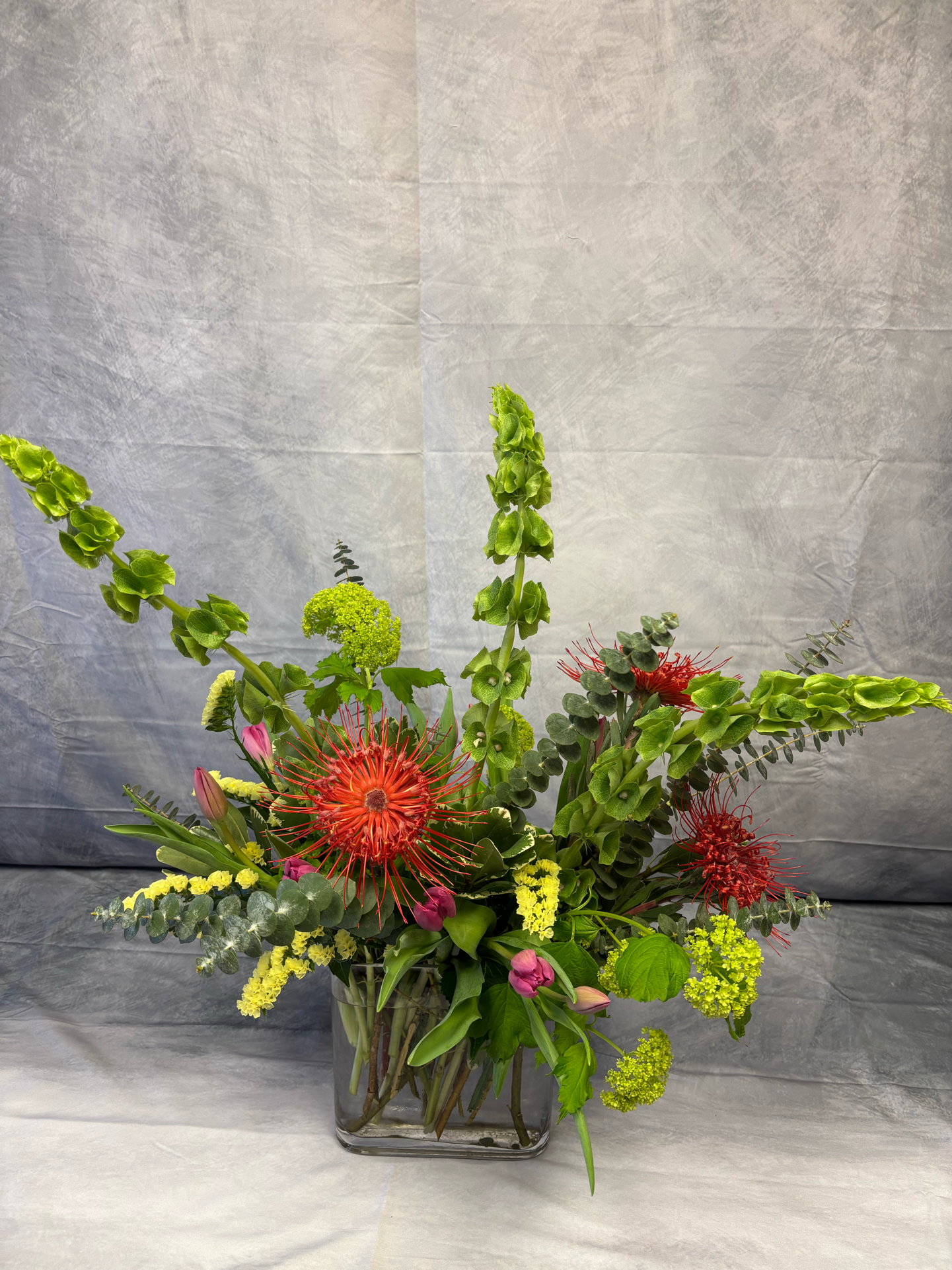 Colorful flower arrangement in a glass vase with various flowers including red pincushion protea, pink tulips, green hydrangeas, and greenery, set against a gray background.