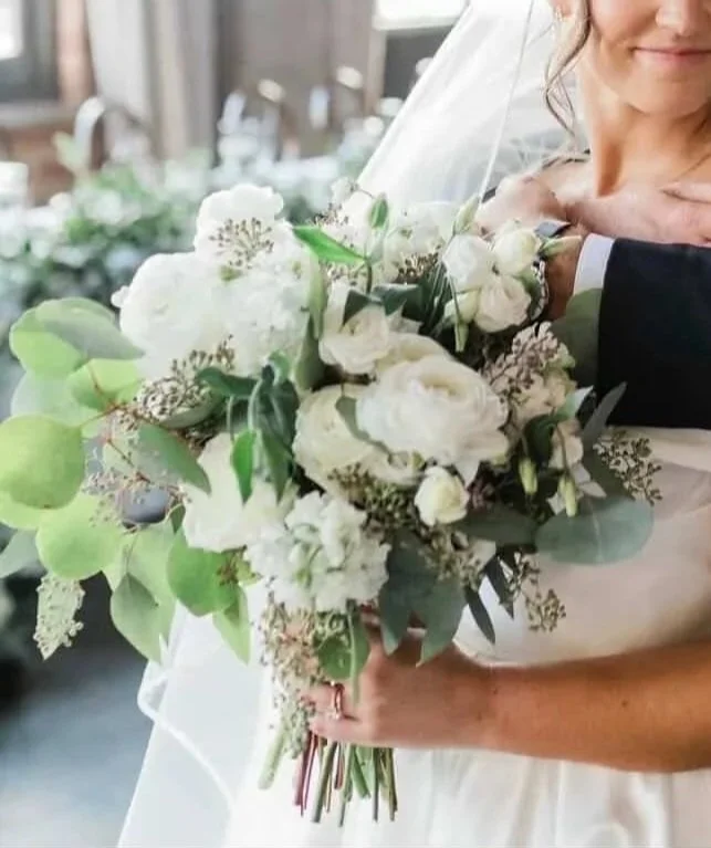 Close-up of a bride holding a bouquet of white flowers, including roses and greenery, at a wedding.