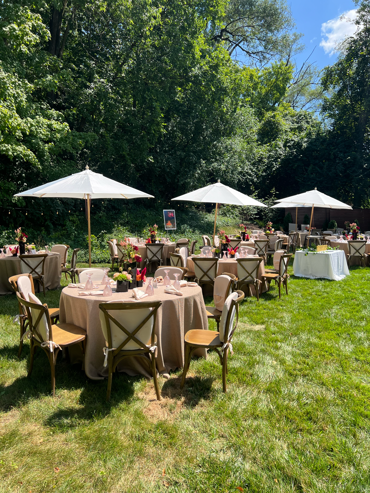 Outdoor event setup with round tables covered with beige tablecloths, decorated with pink and red flower centerpieces, glasses, and napkins. White umbrellas shading the tables are set on a grassy lawn, surrounded by trees.