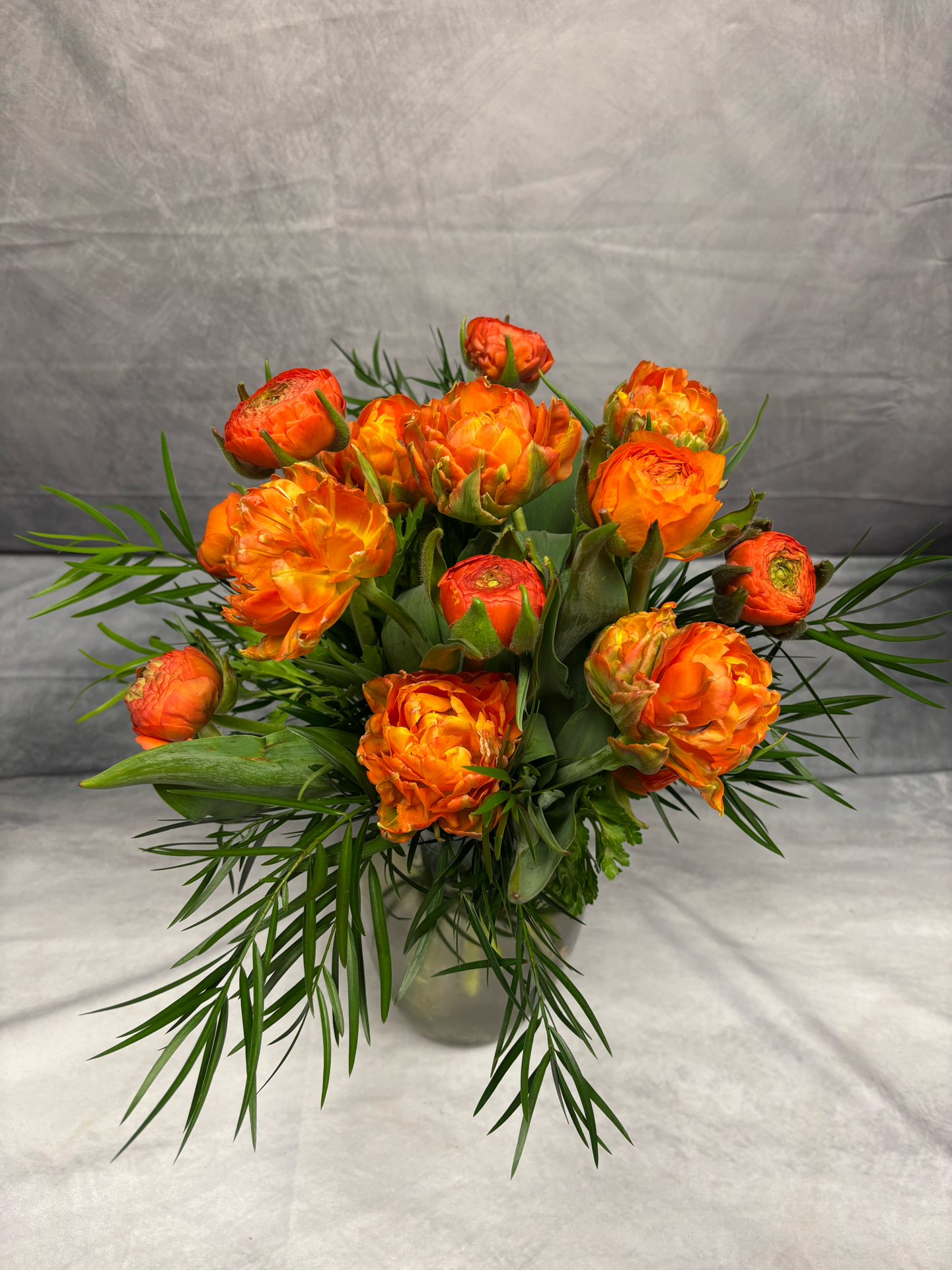 A bouquet of orange double tulips with lush green foliage arranged in a glass vase on a white surface with a gray textured background.