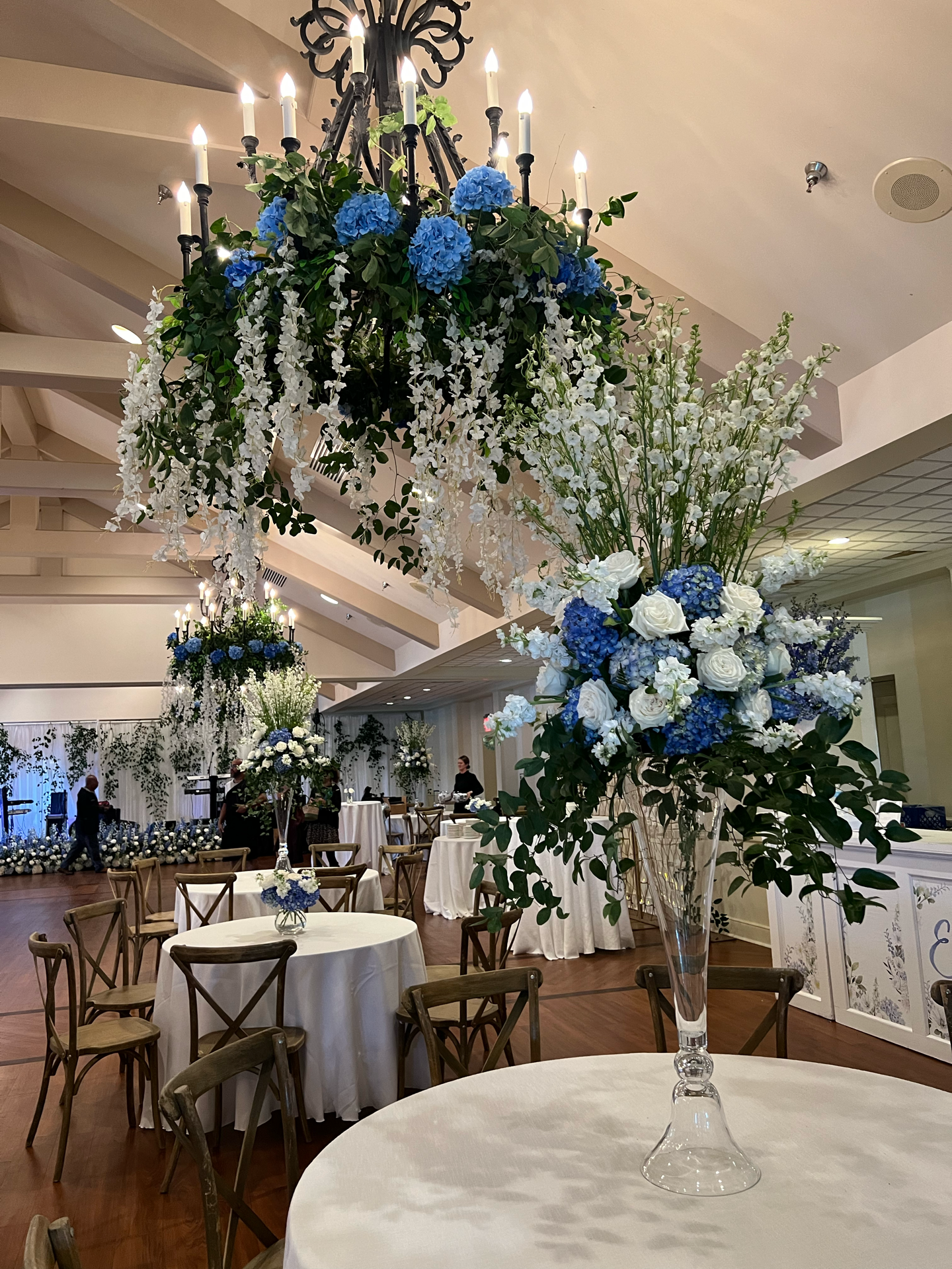 A large floral centerpiece with blue hydrangeas, white roses, and white flowers in a tall glass vase, placed on a round table with a white tablecloth in a decorated event space.