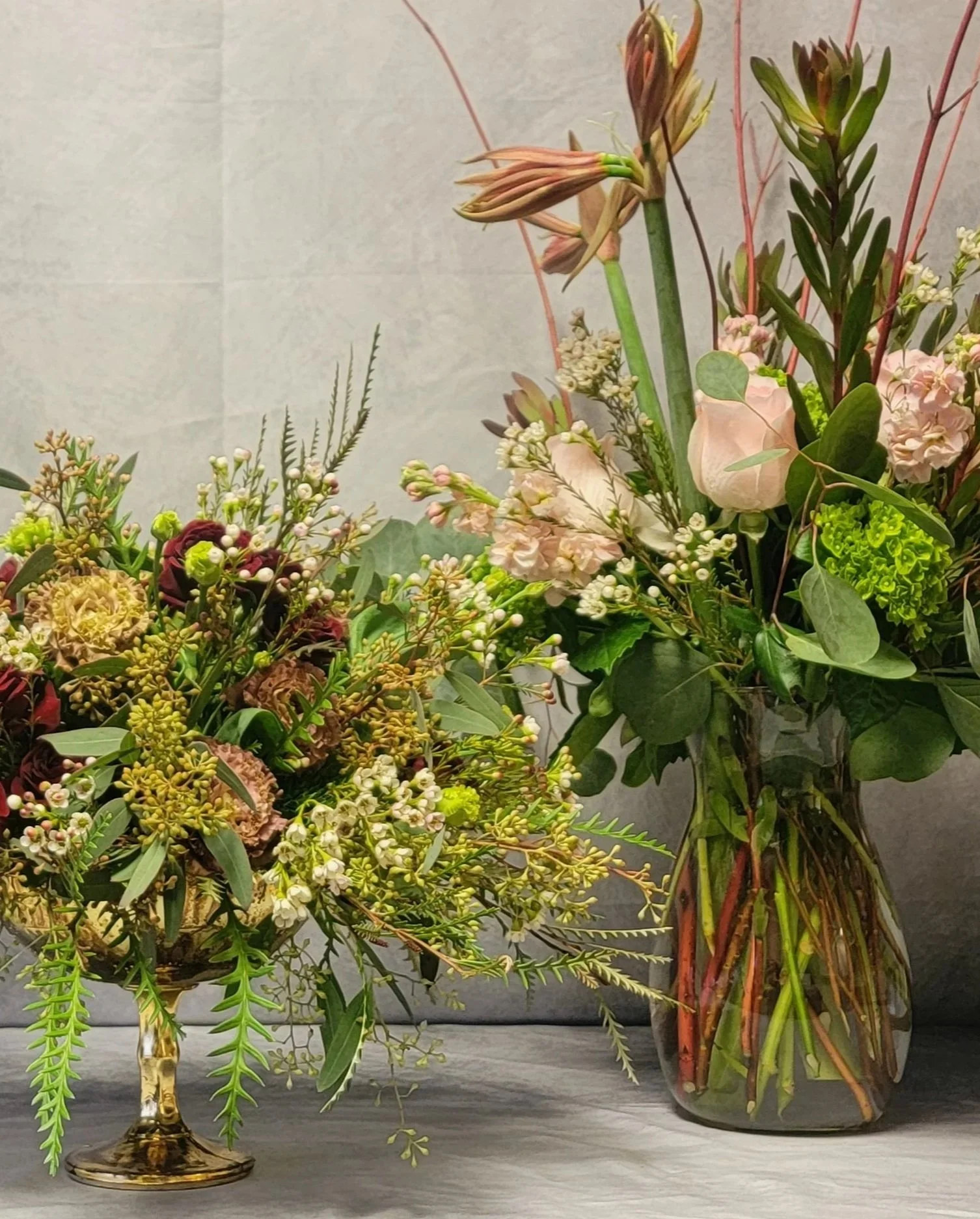 Two floral arrangements with pink, cream, and green flowers and foliage, one in a gold pedestal bowl and the other in a clear glass vase, placed on a gray surface against a neutral background.