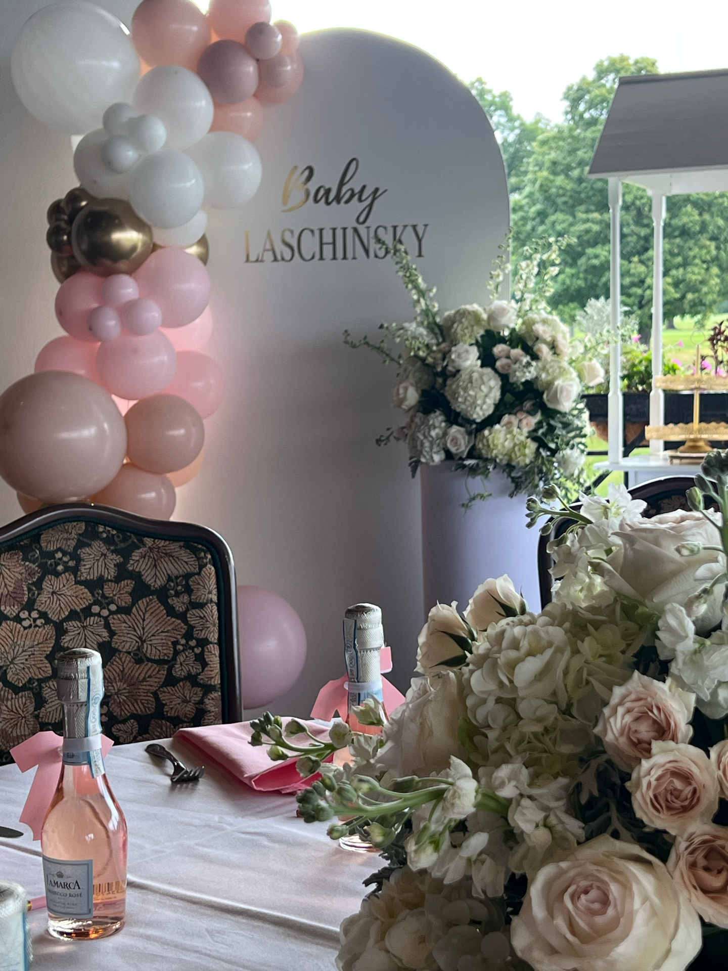 Decorations for a baby shower, featuring pink, white, gold, and gray balloons, a large floral arrangement with white and blush roses, a table with pink napkins, bottles of rosé wine, and a white backdrop with the text "Baby LASCHINSKY."
