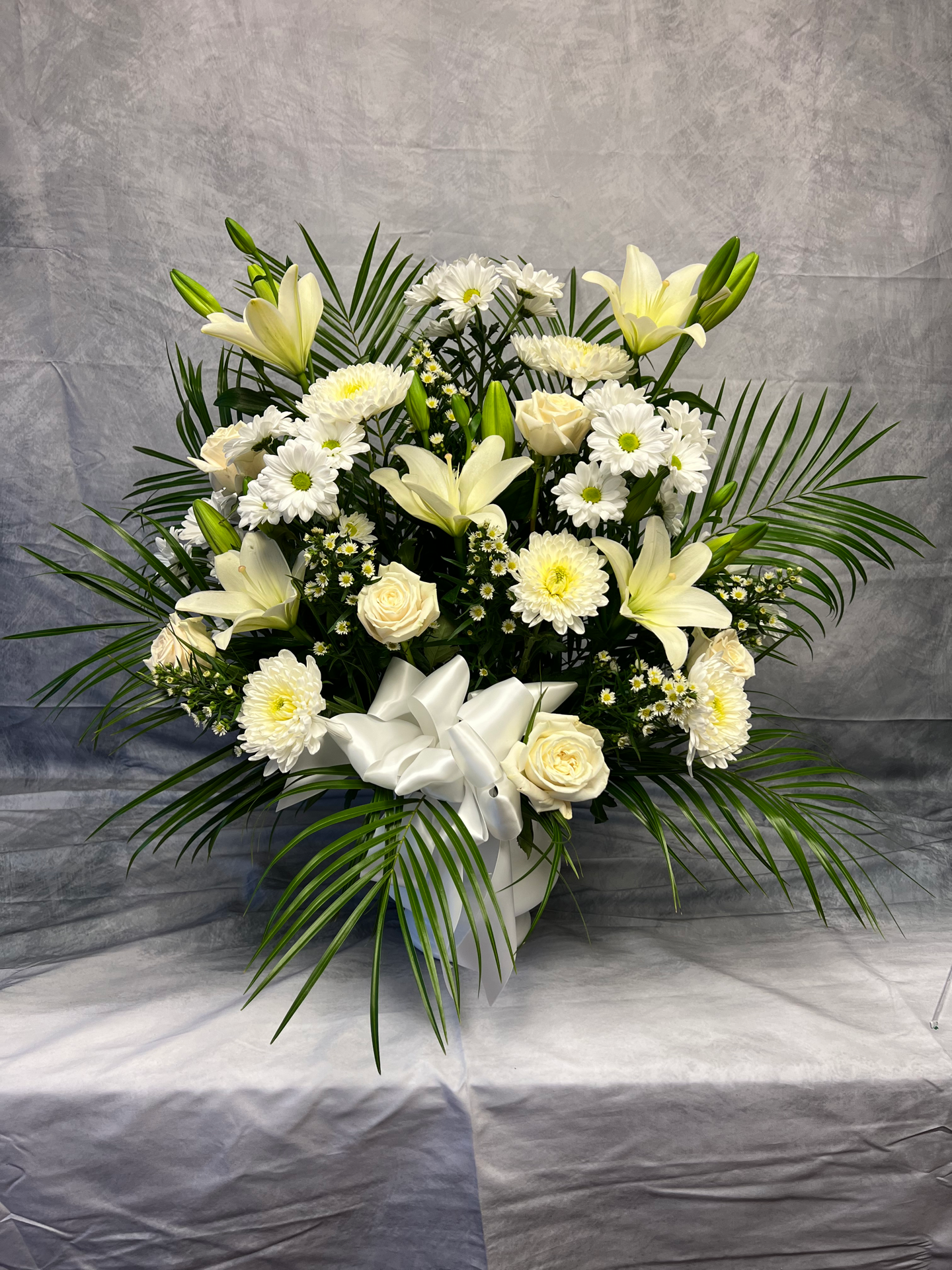 A white floral arrangement with lilies, roses, daisies, and greenery in a white container.