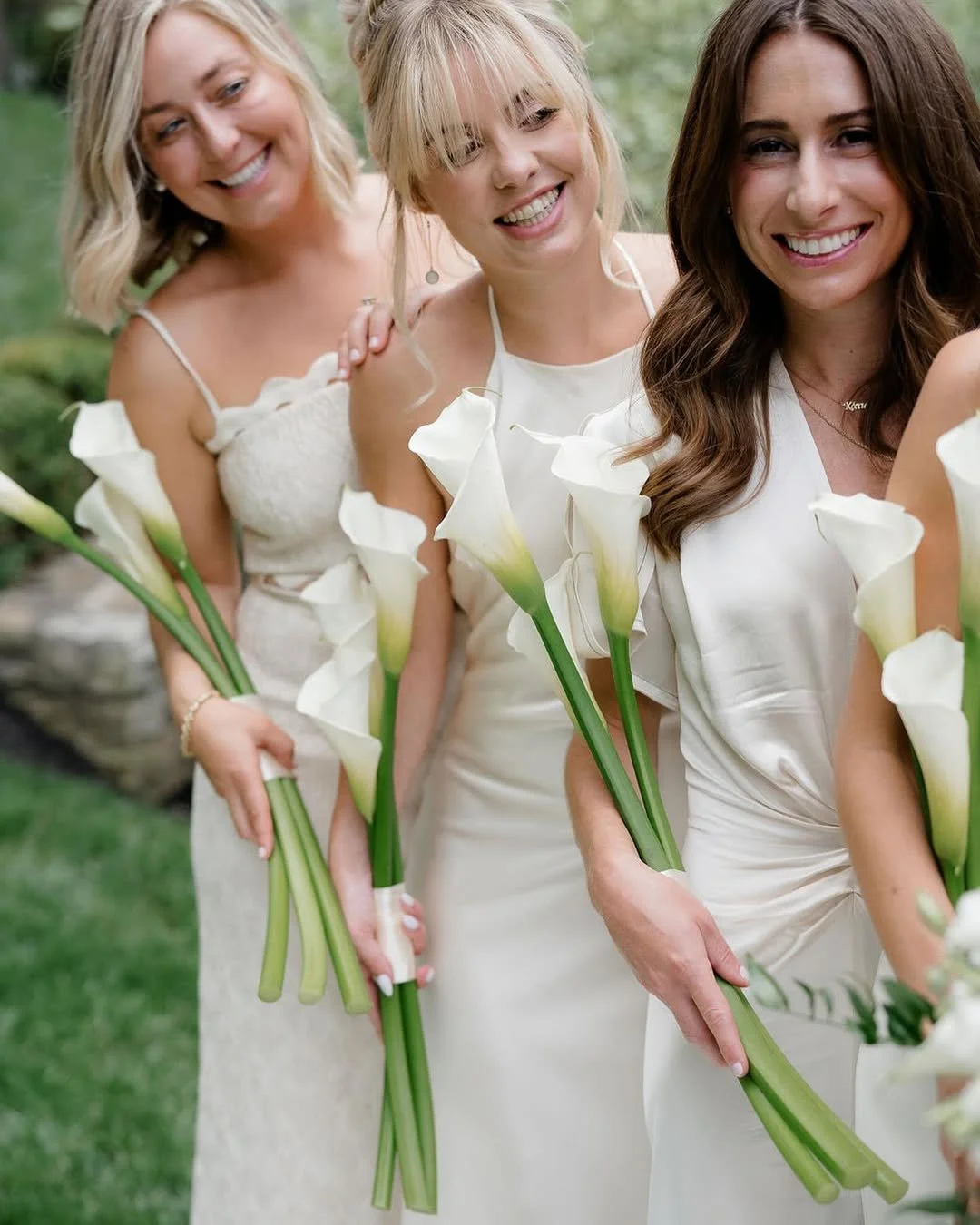 Three women dressed in white holding white calla lily flowers outdoors, smiling and posing for the photo.