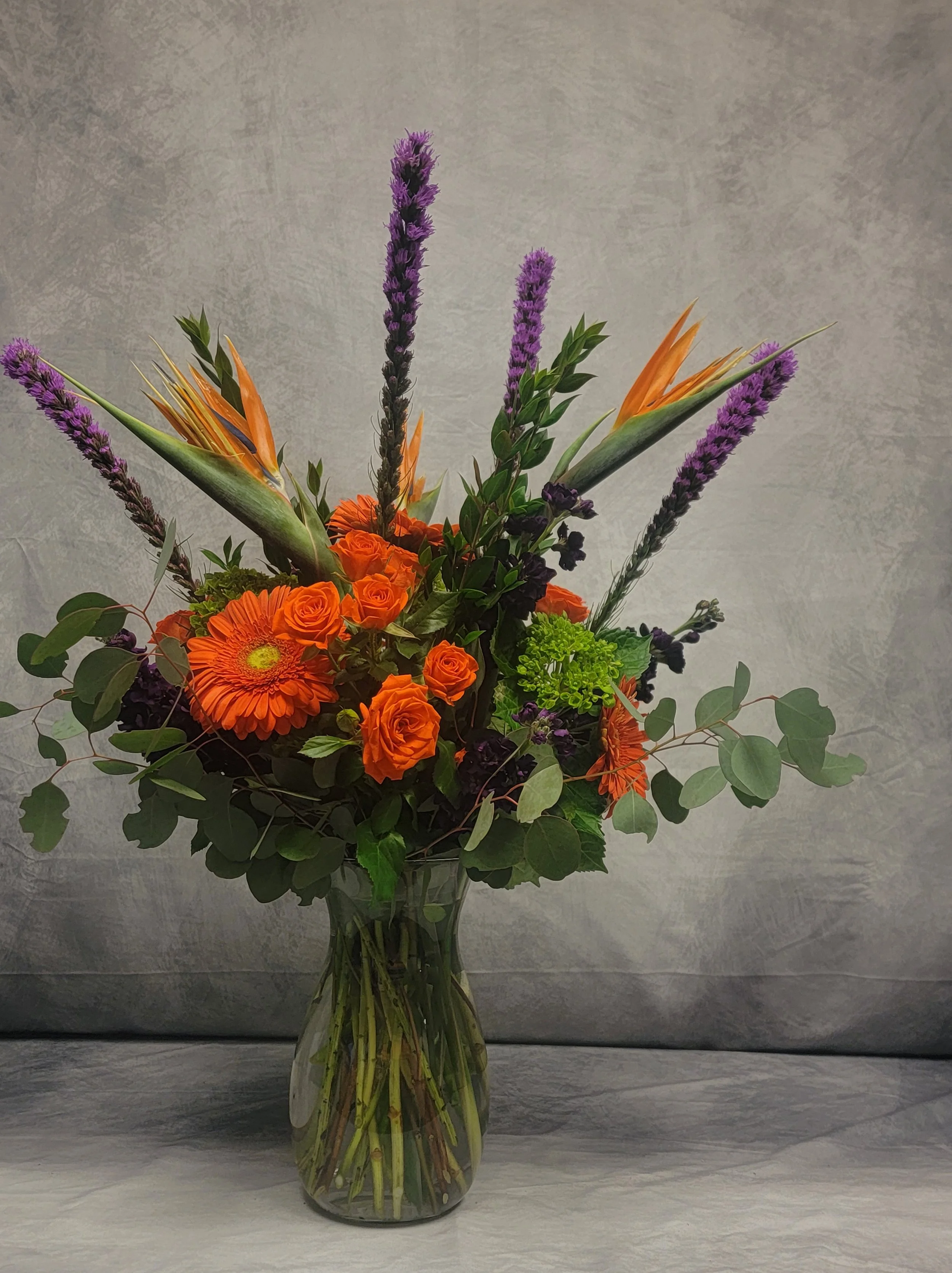 A bouquet with orange roses, gerbera daisies, bird of paradise flowers, purple lavender, and green foliage in a glass vase.